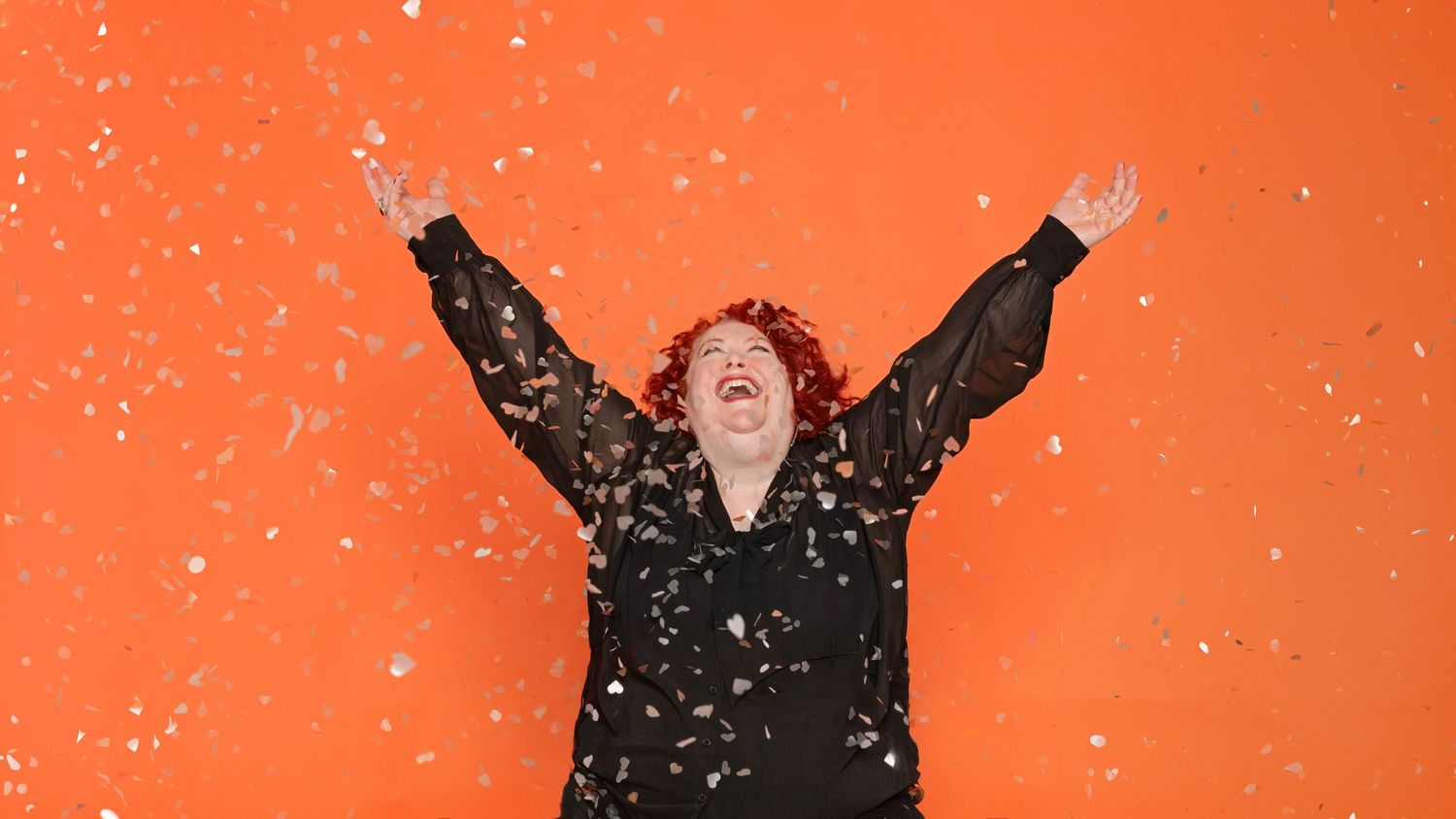 Joyful woman with curly red hair laughs with arms raised as confetti falls around her against a bright orange background.