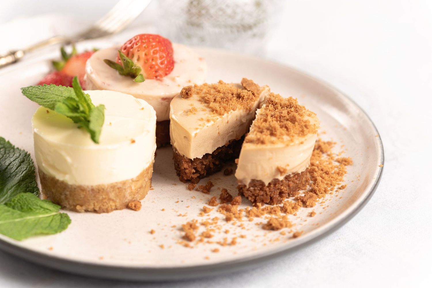 Mini cheesecakes with graham cracker crust, topped with cream cheese filling, mint leaves, and fresh strawberry on a white plate.