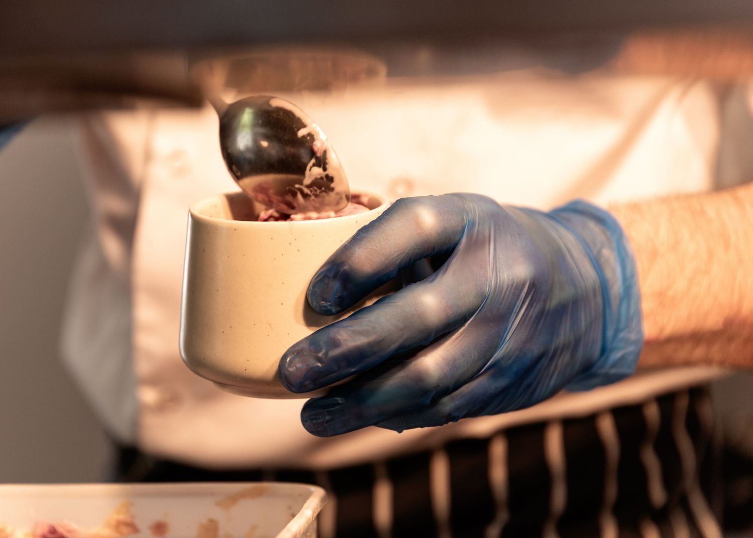 A gloved hand holds a cup of chocolate ice cream being scooped at an ice cream shop.
