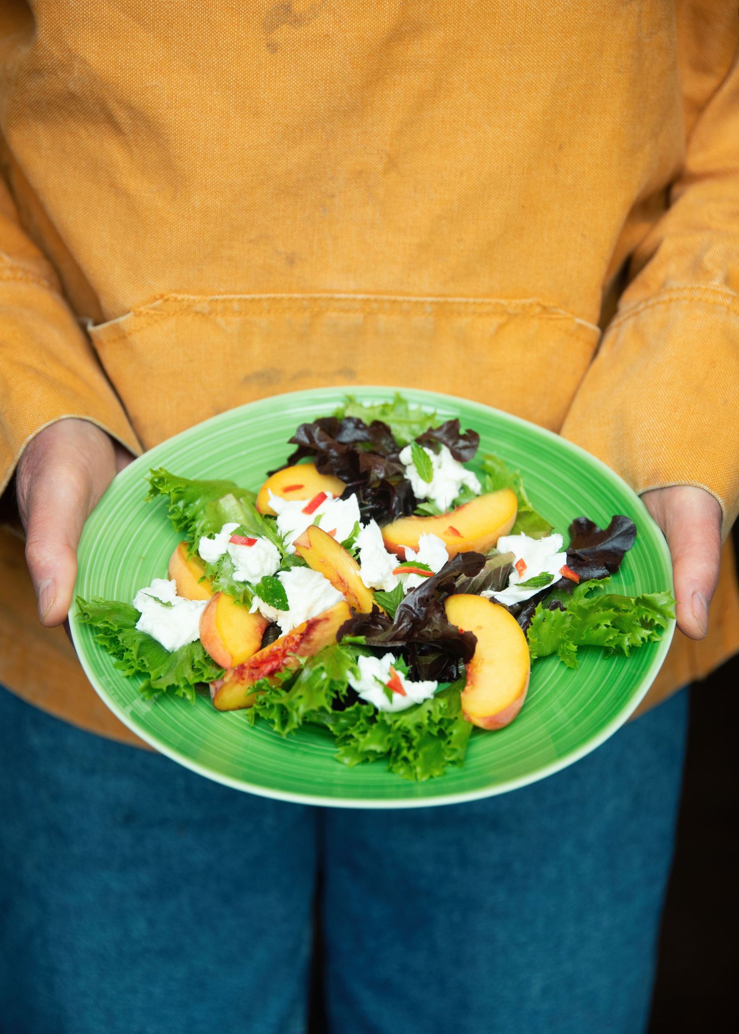 Person in orange top holding a green plate with fresh salad featuring peach slices, white cheese, and mixed greens.