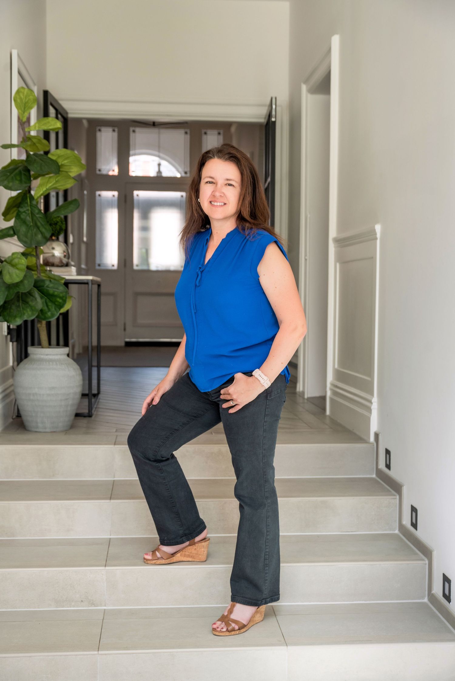 Woman in blue sleeveless top and dark pants posing on stairs in elegant home entryway.