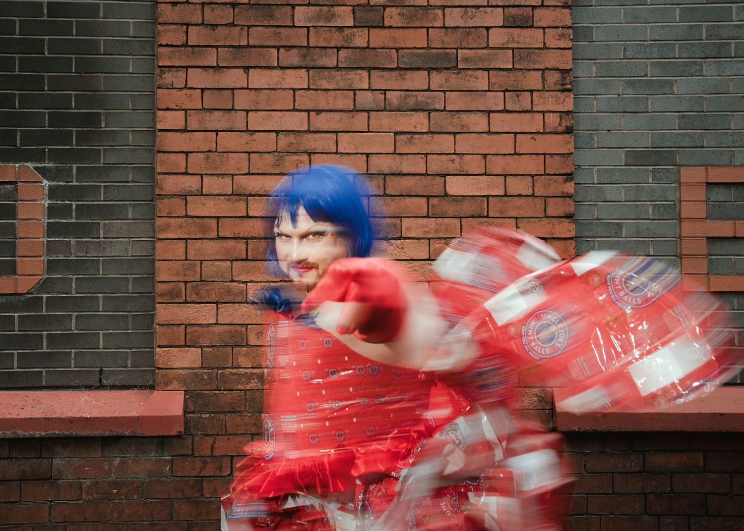 Woman in blue wig and red dress spinning energetically against a brick wall backdrop.