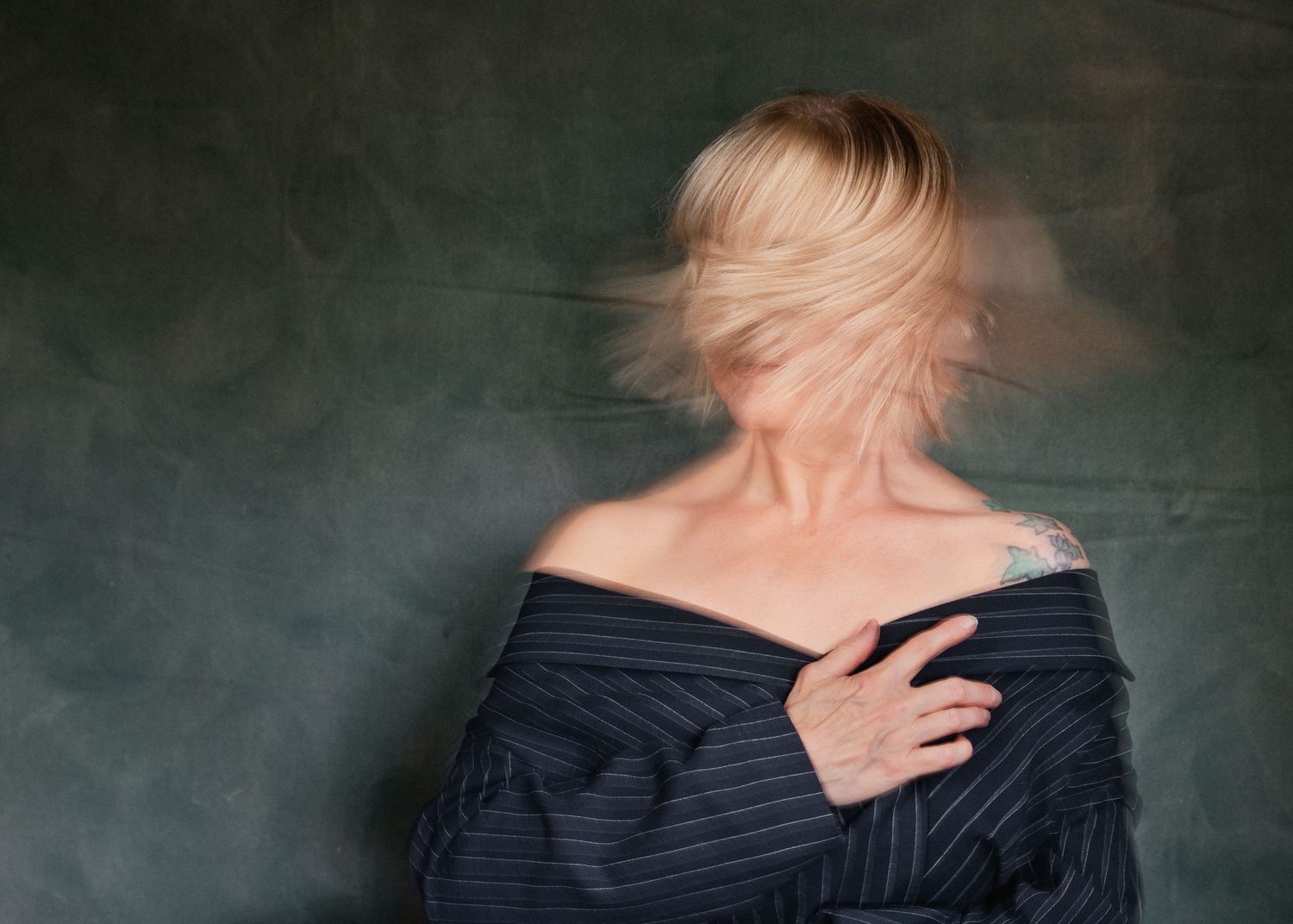 Blonde woman in off-shoulder pinstripe blazer with hair blowing across face, artistic motion blur portrait.