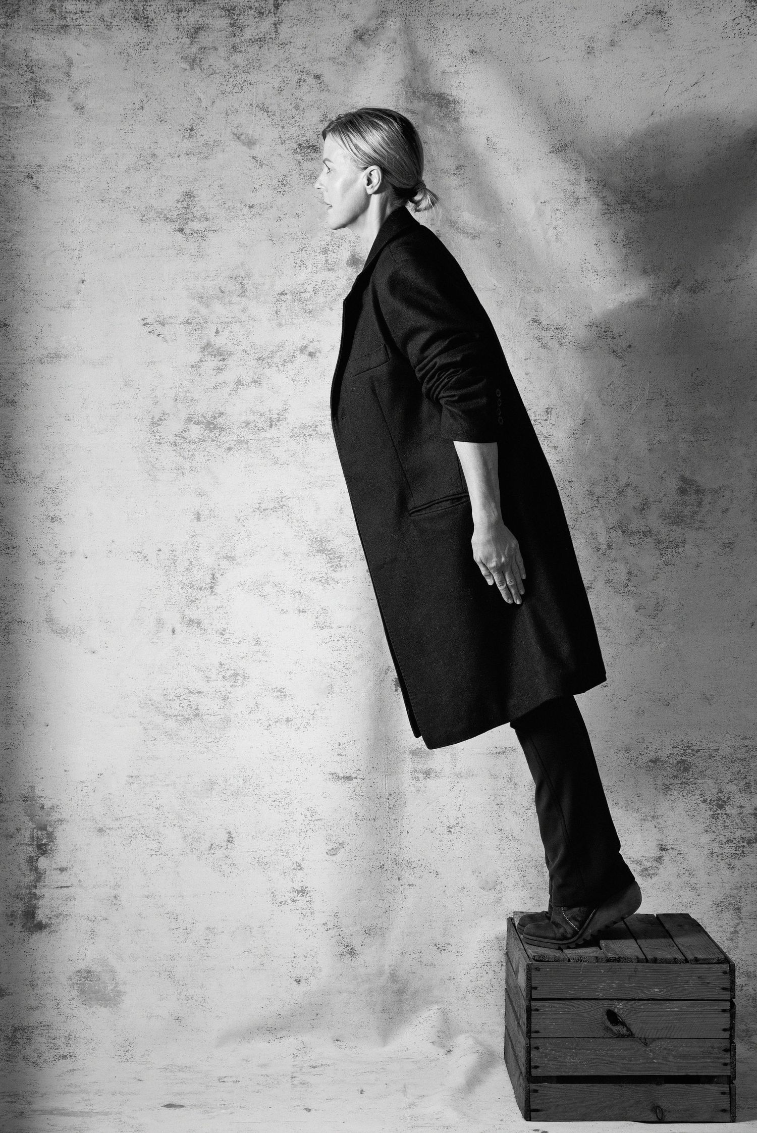 Black and white portrait of a woman in a long dark coat standing on a wooden crate against a textured wall.