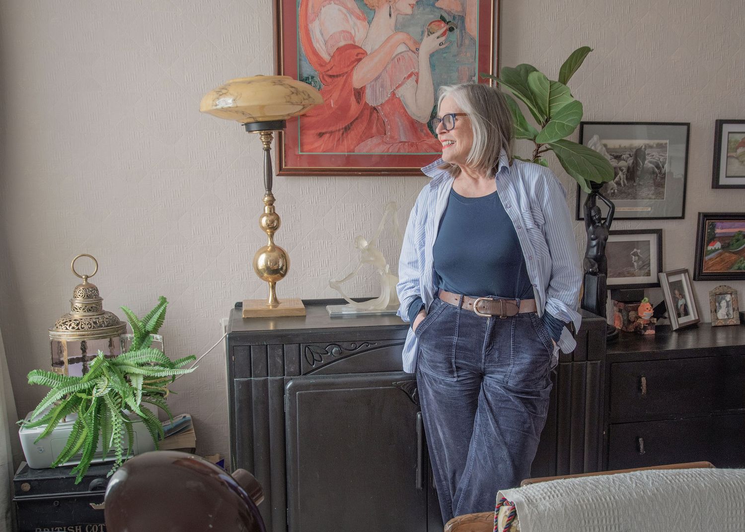 Older woman in blue jeans and cardigan standing in eclectic home interior with vintage lamp and artwork.