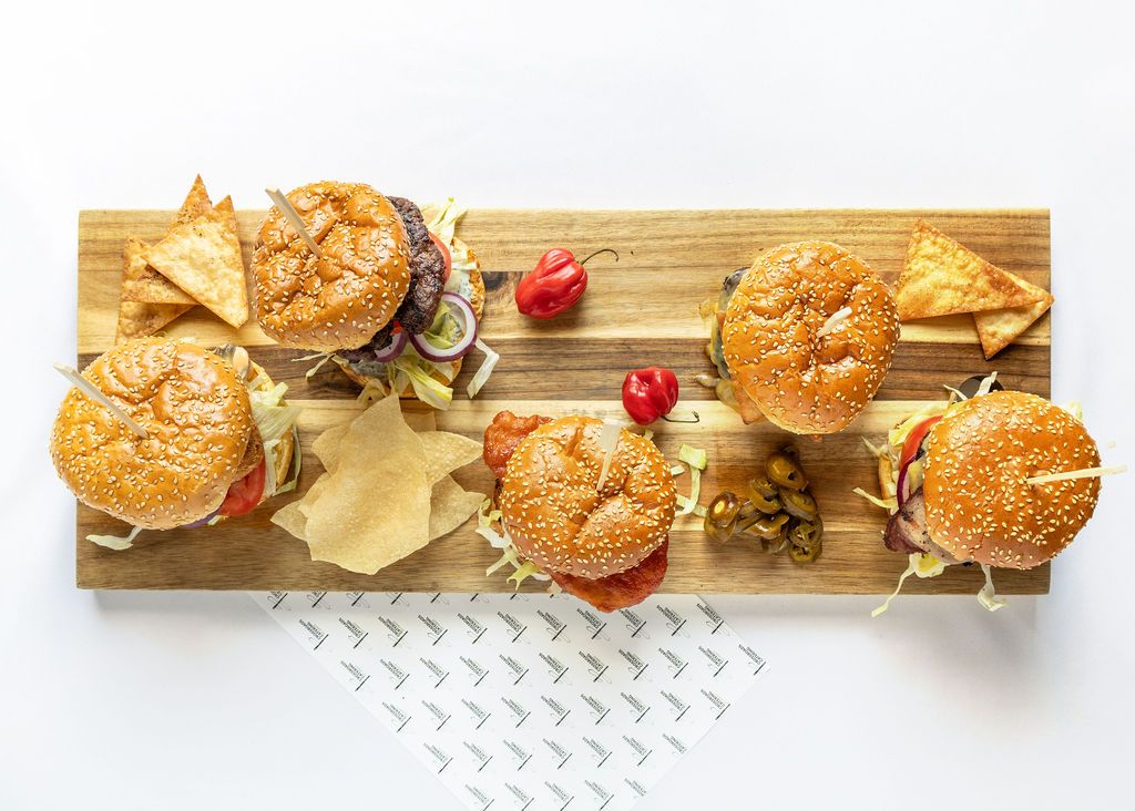 Six gourmet burgers on a wooden serving board with tortilla chips and fresh peppers on a white background.