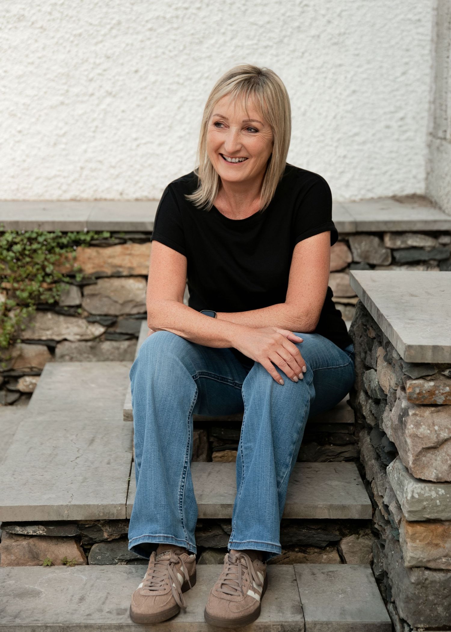 Smiling middle-aged woman with blonde hair sitting on stone steps wearing black top and jeans.