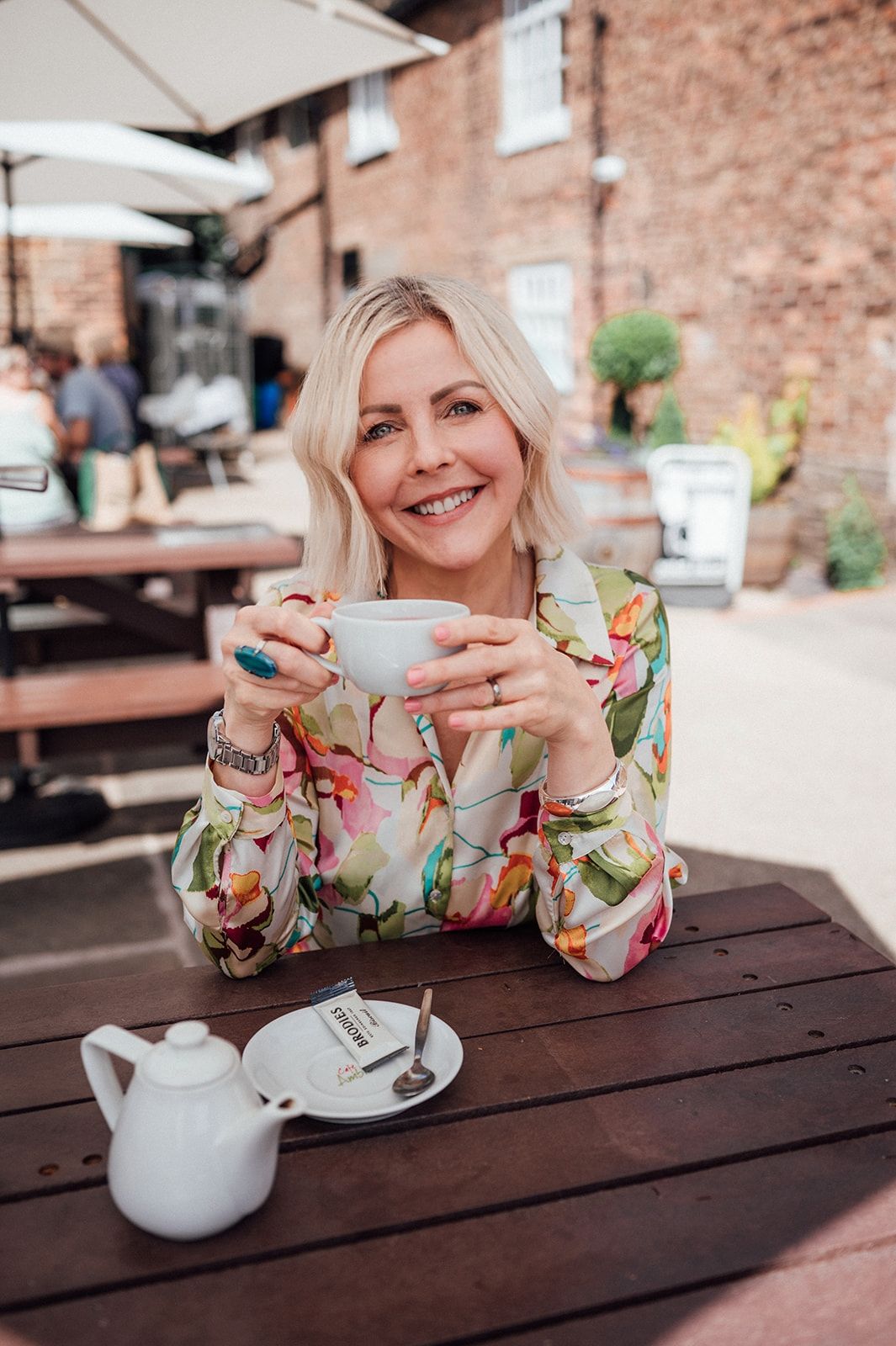 Smiling blonde woman in floral blouse holding tea cup at outdoor cafe table with white teapot.