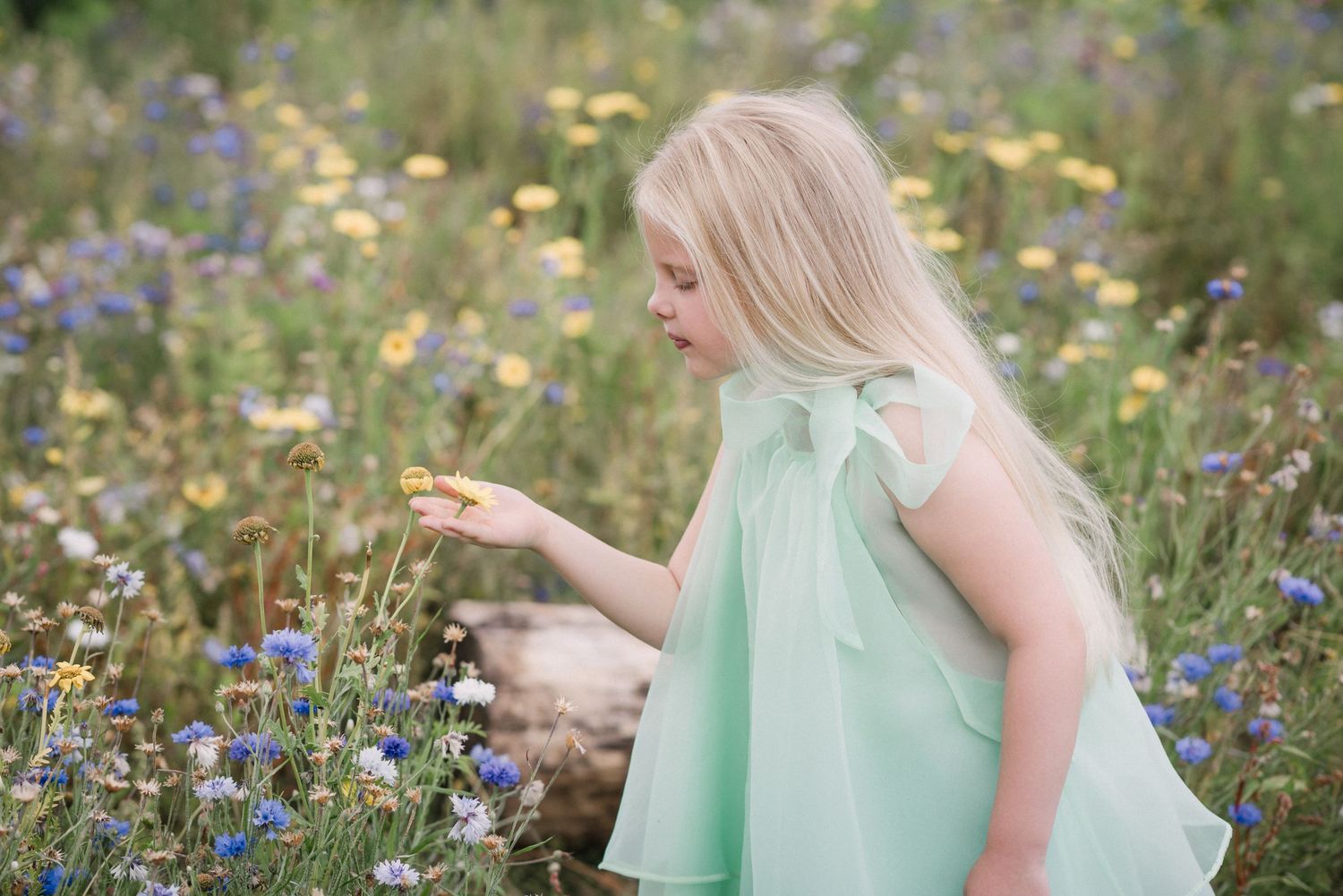 Young blonde girl in mint dress reaching for wildflowers in a colorful meadow field.