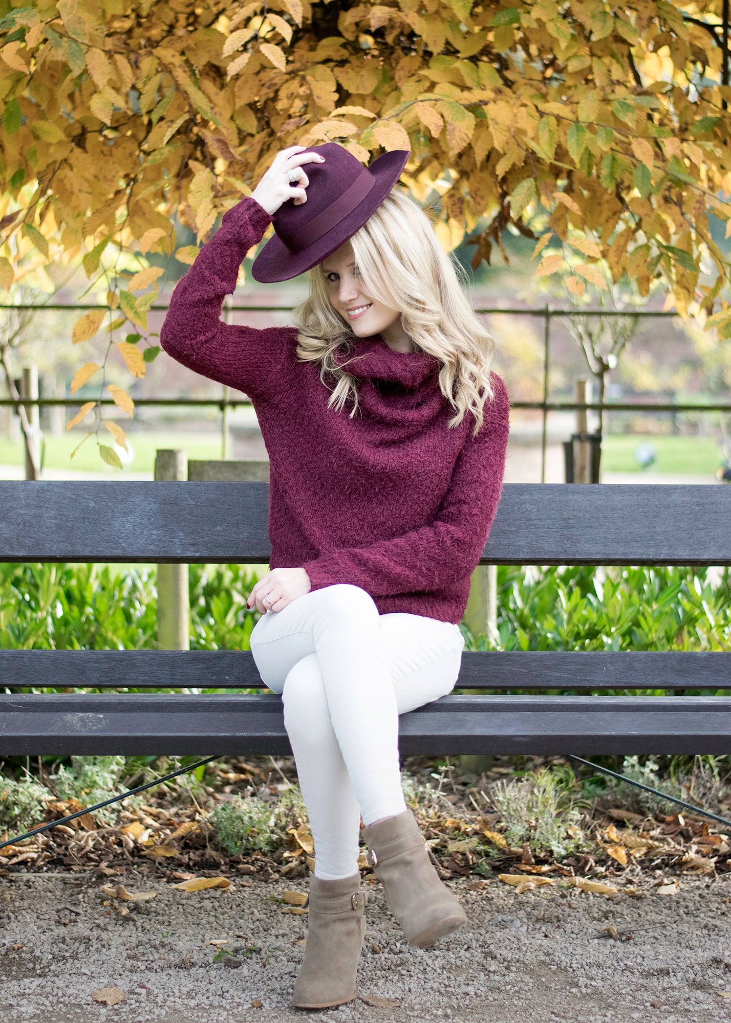Blonde woman in burgundy sweater, white jeans, and tan boots sits on park bench wearing matching hat.
