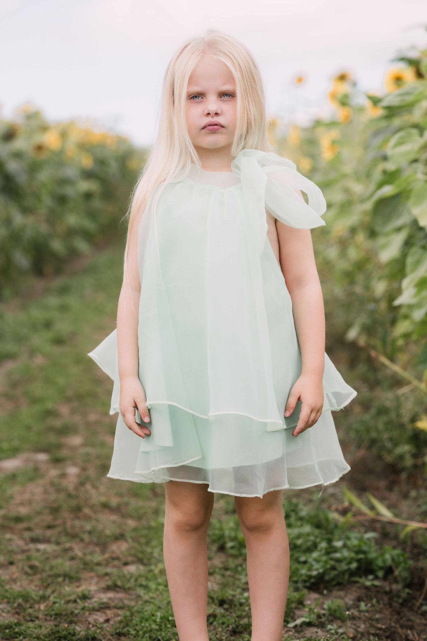 A toddler in a mint green chiffon dress stands in a garden setting with sunlight filtering through foliage.