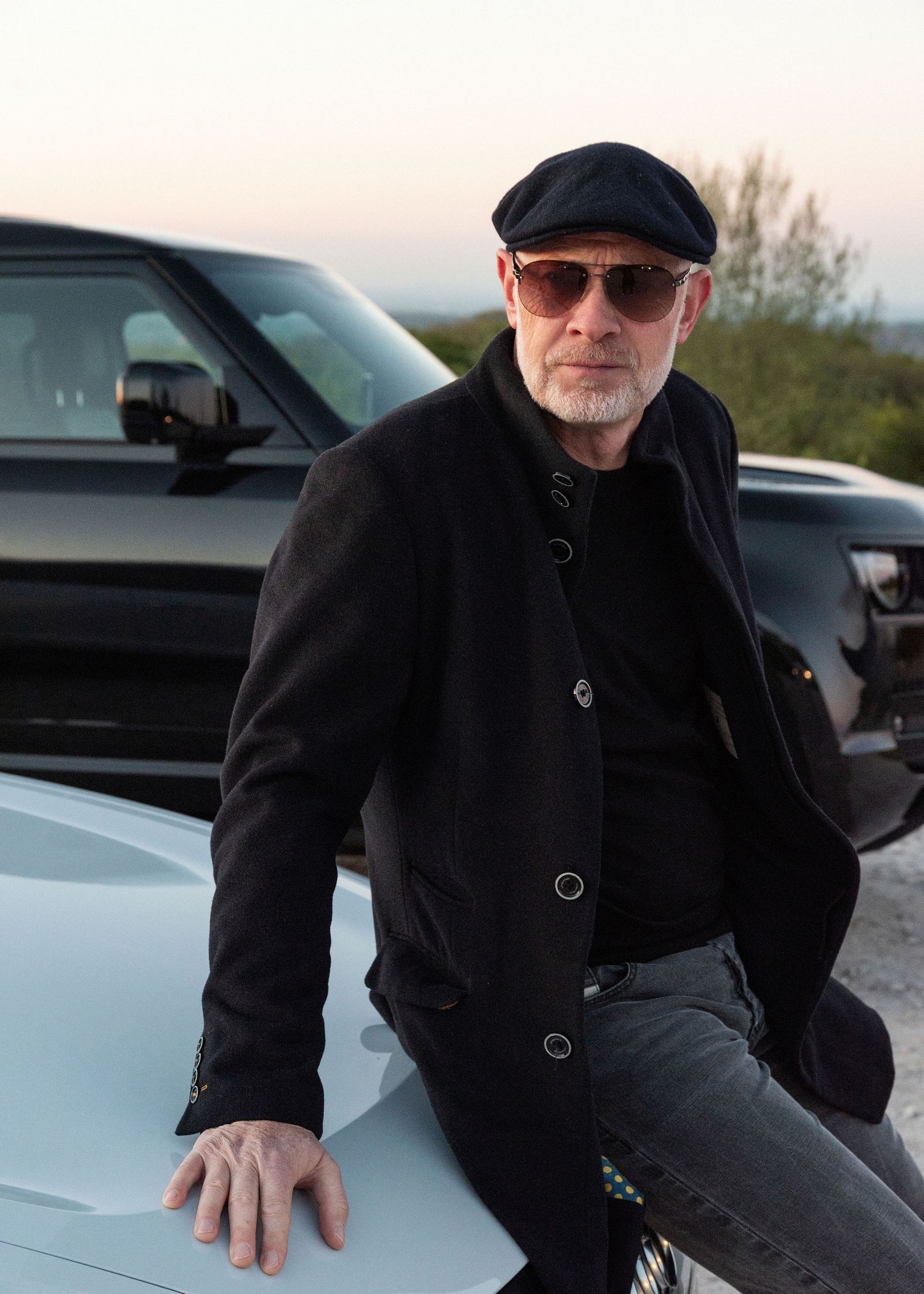 Middle-aged man in black cap, sunglasses, and jacket leaning on a white car outdoors at dusk.