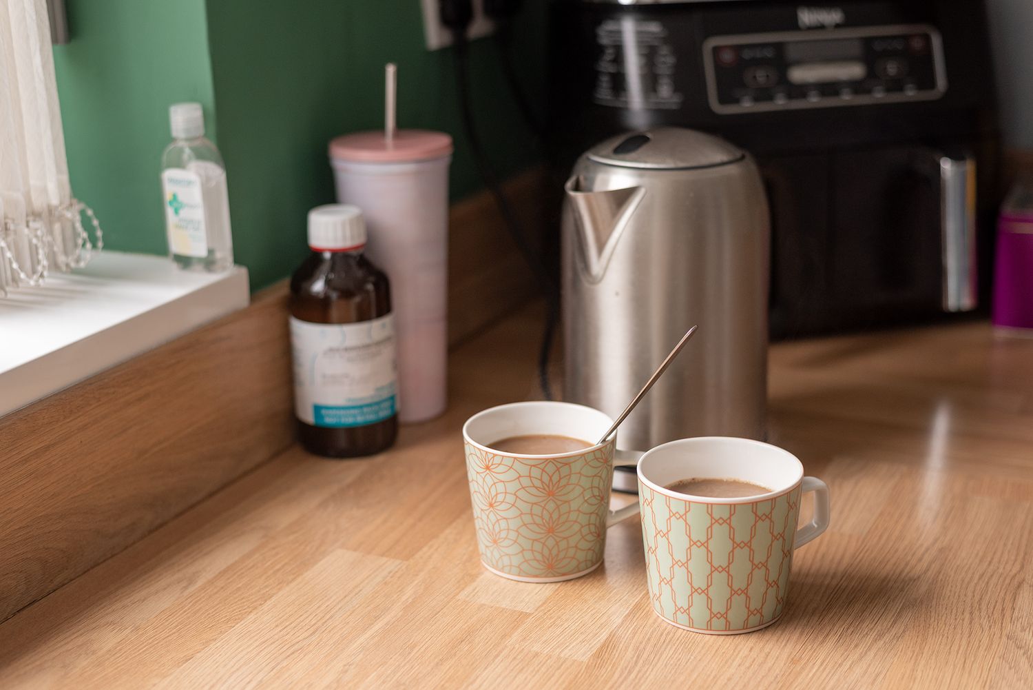 Two ceramic mugs with hot drinks beside a silver kettle on a wooden countertop with a coffee machine in background.