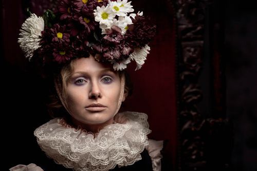 Woman in Victorian costume with floral crown and lace ruff collar poses dramatically in dark, moody setting.