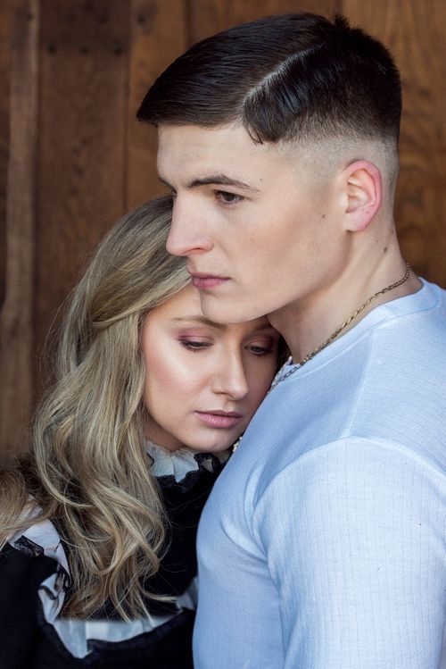 Young couple embracing tenderly, man in blue shirt holds blonde woman close against rustic wood background.