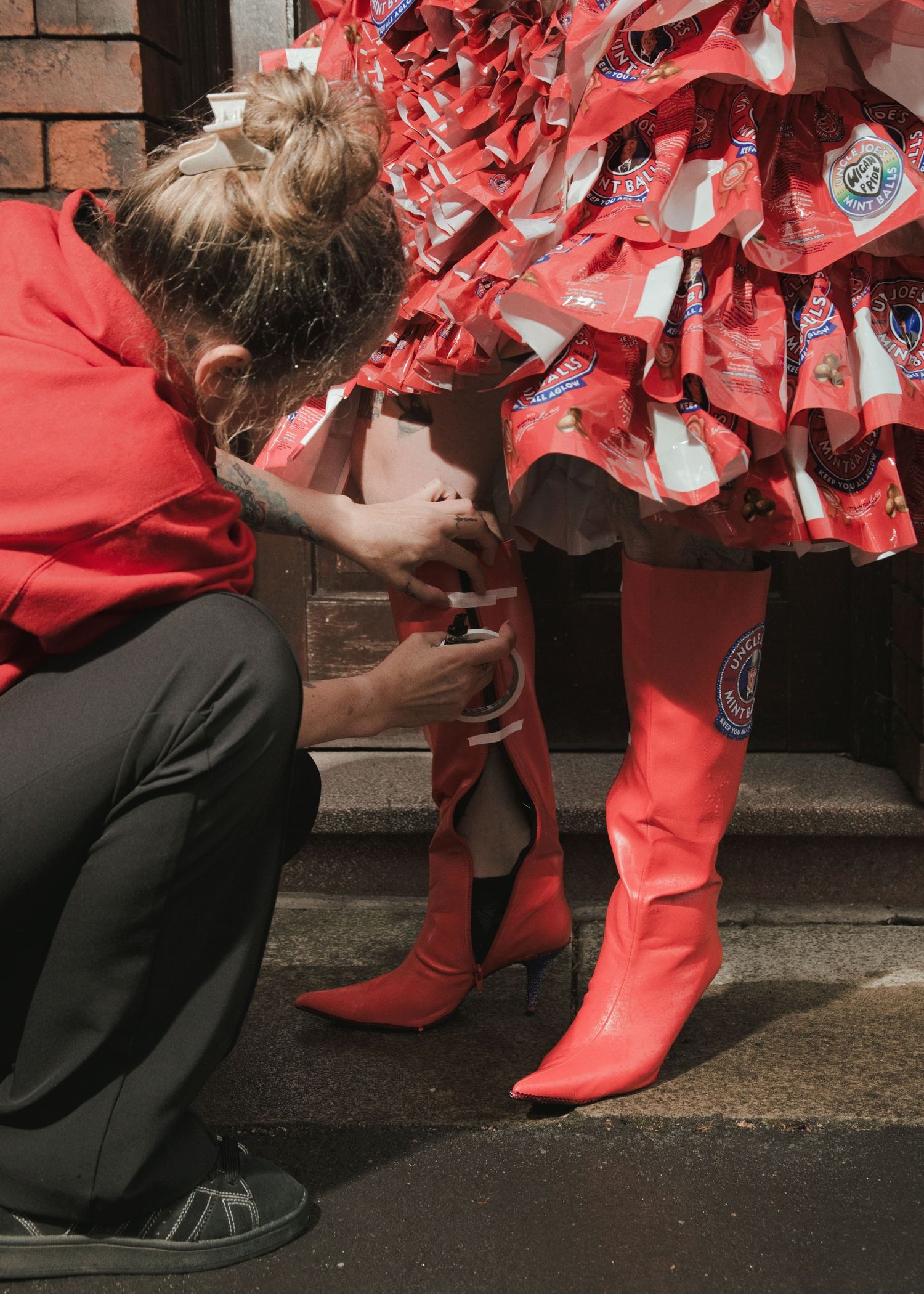 Person in red adjusting bright red pointed boots on model wearing a skirt made of red chip packets.