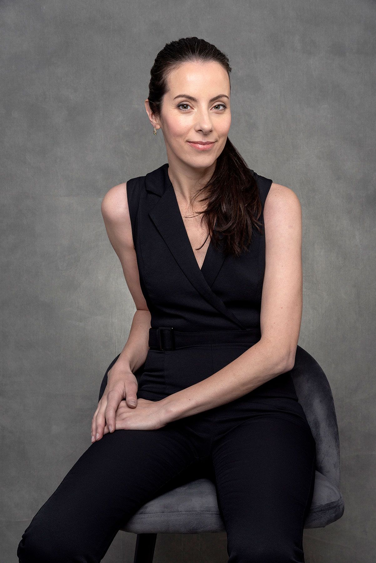 Woman in black sleeveless jumpsuit seated on gray chair, smiling confidently against neutral studio background.