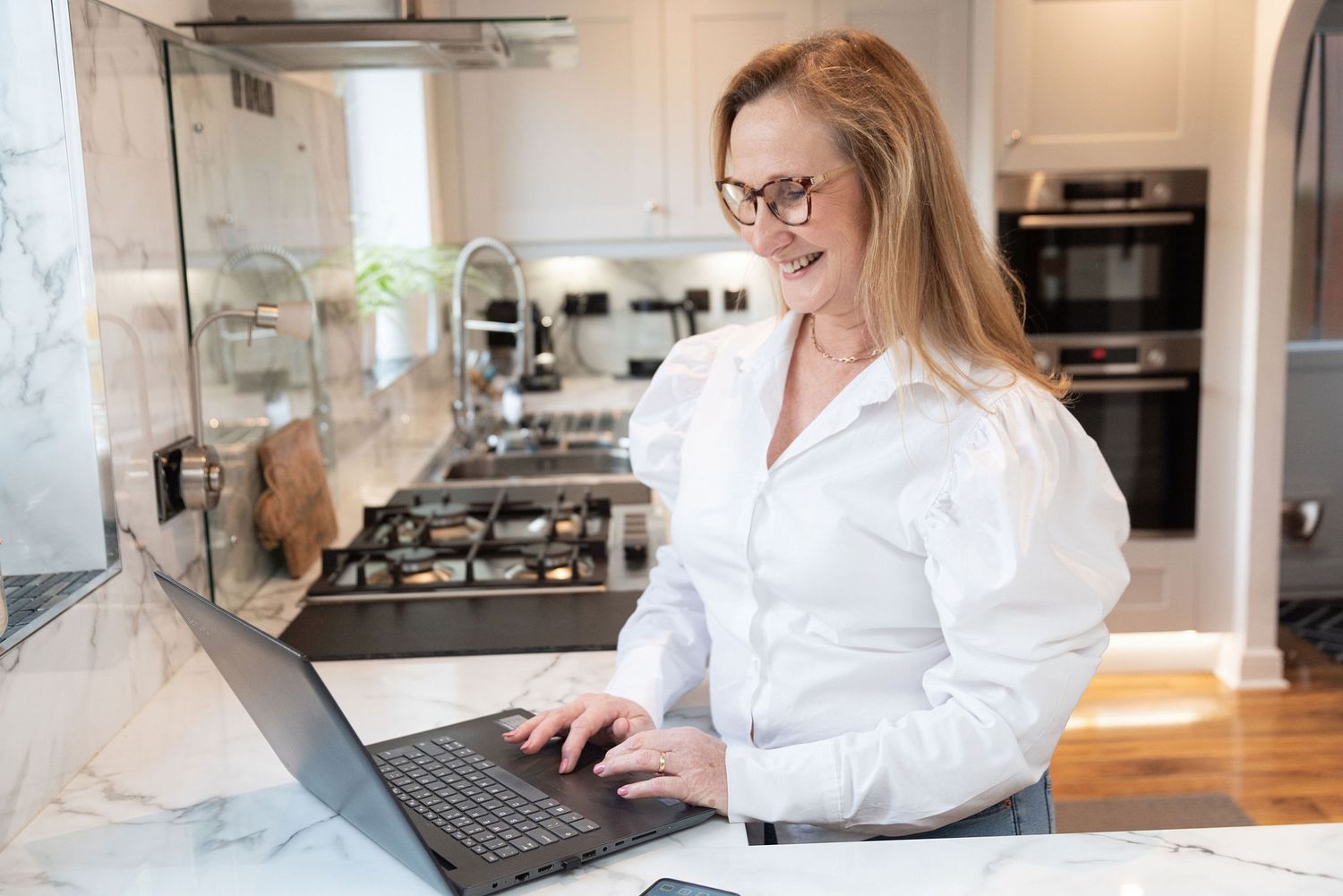 Person typing on laptop while standing at kitchen counter with marble backsplash.