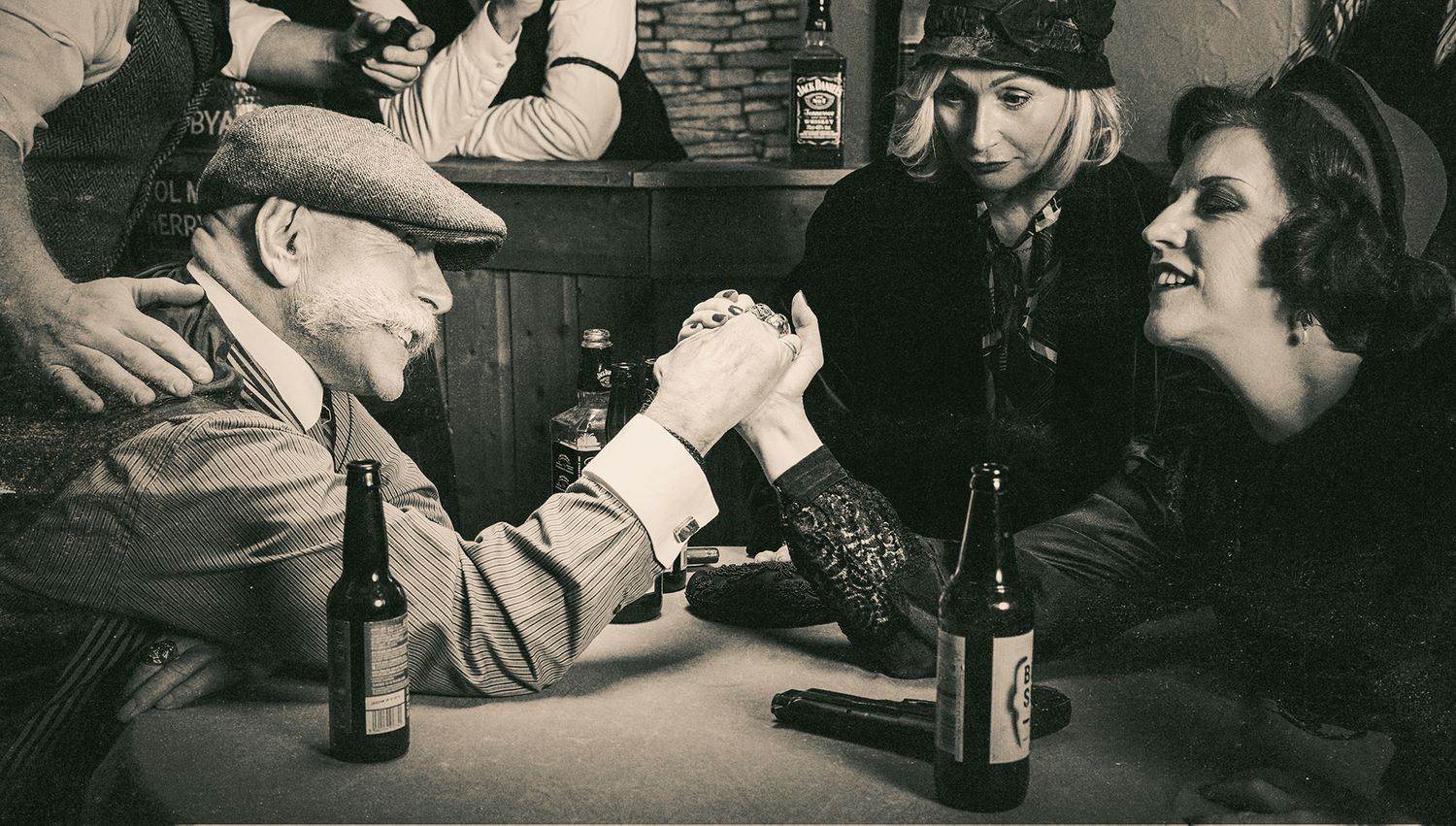 Vintage black and white photo of people arm wrestling at a bar counter with beer bottles.
