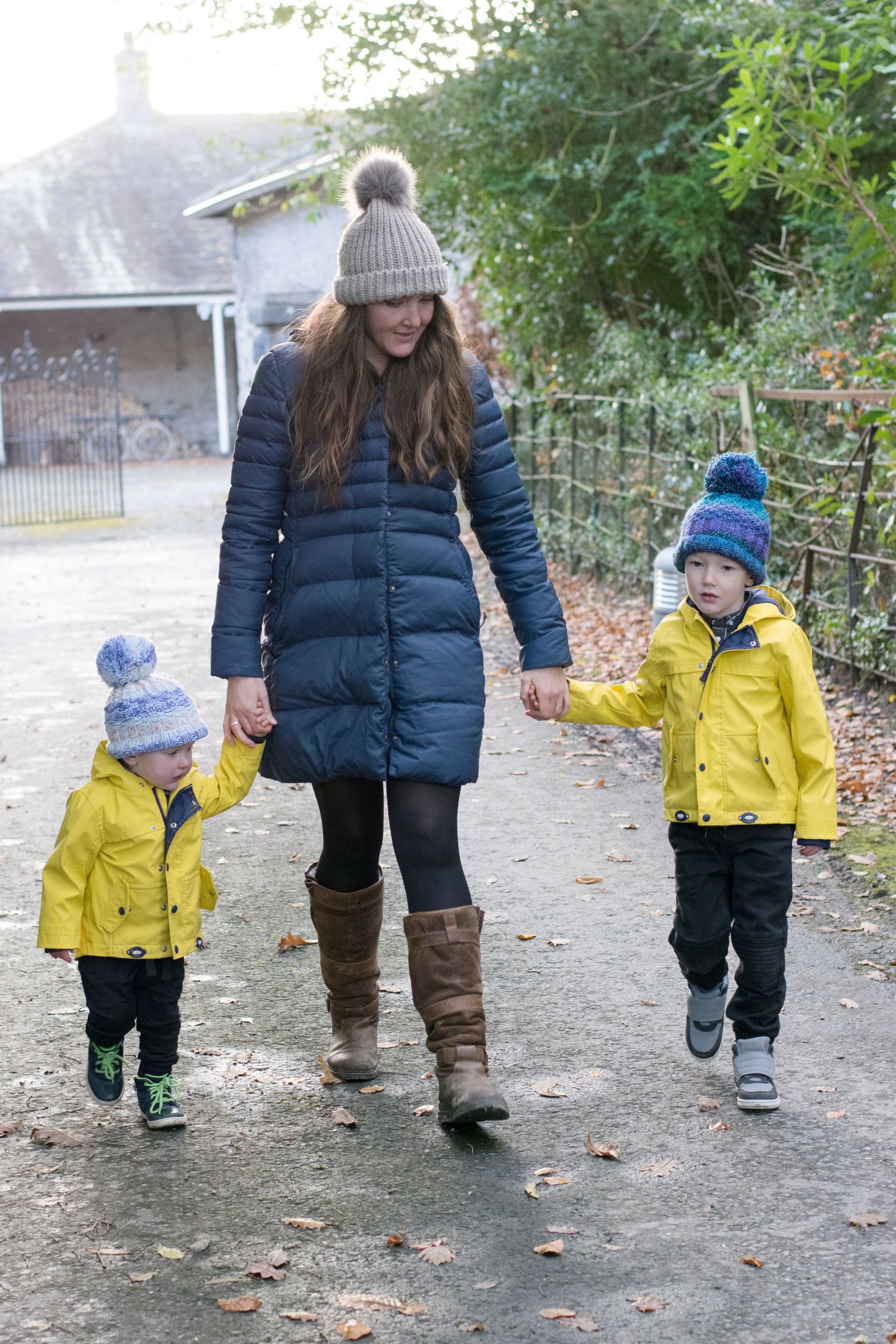 Parent and toddler in yellow raincoat walking together on autumn path with fallen leaves.
