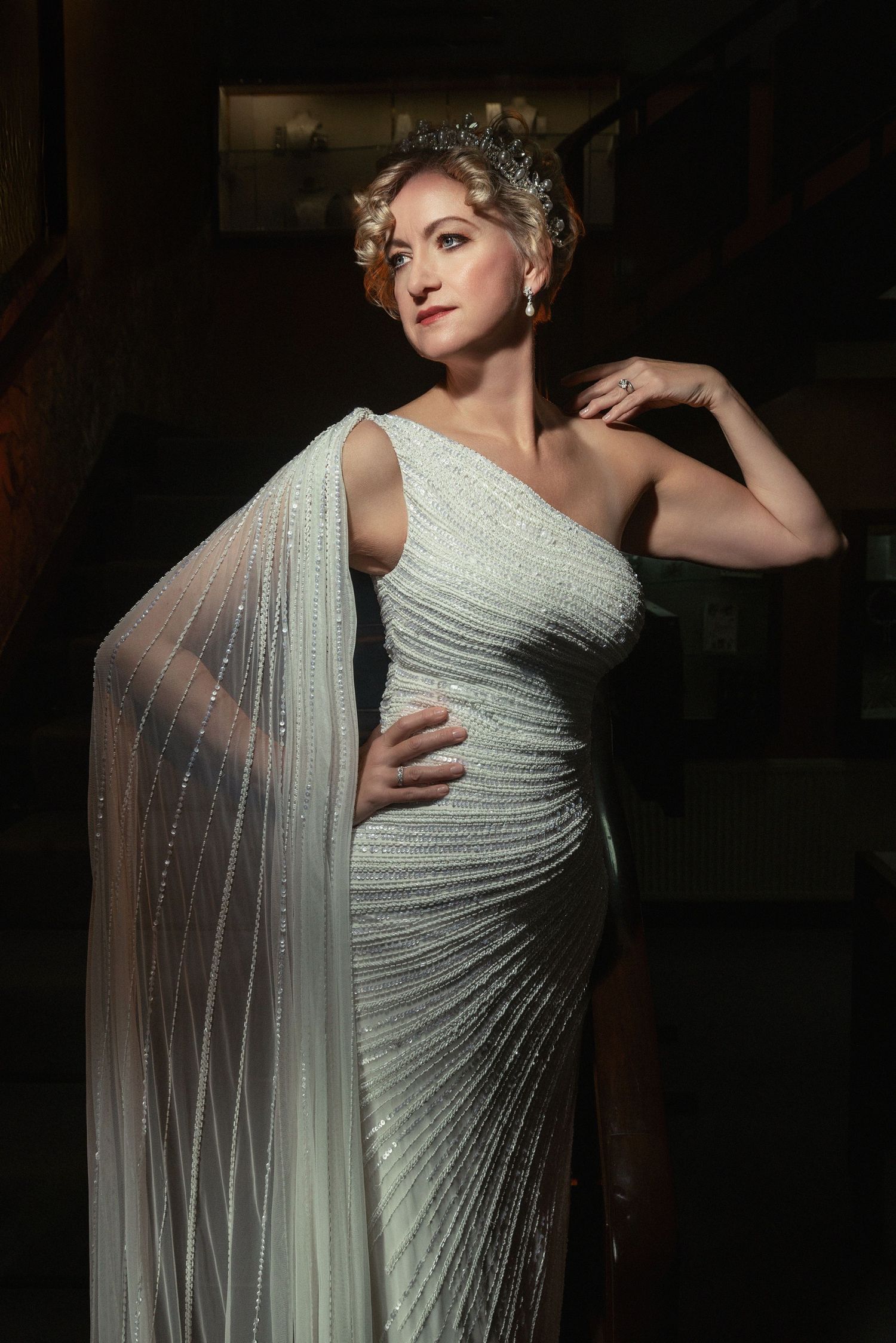 Elegant portrait in a beaded one-shoulder gown with flowing cape and crystal headpiece against dark backdrop.