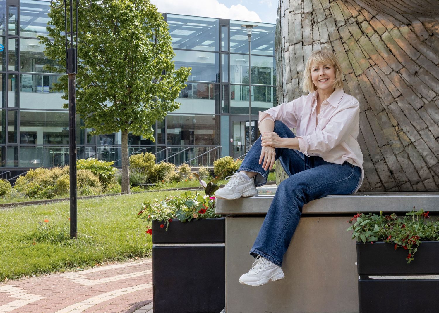 Woman in pink blouse and jeans sits relaxed on modern planter outside contemporary glass office building.