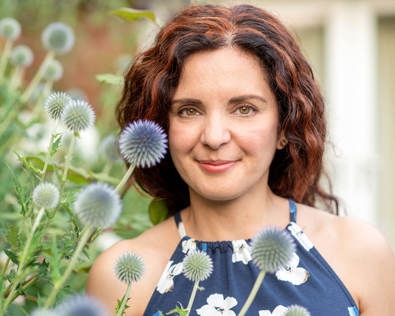 Woman with curly red hair smiling among globe thistle flowers in a lush garden setting.