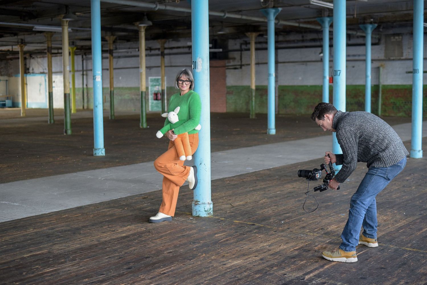 Photographer captures model in orange pants and green top posing by teal pillar in empty warehouse.