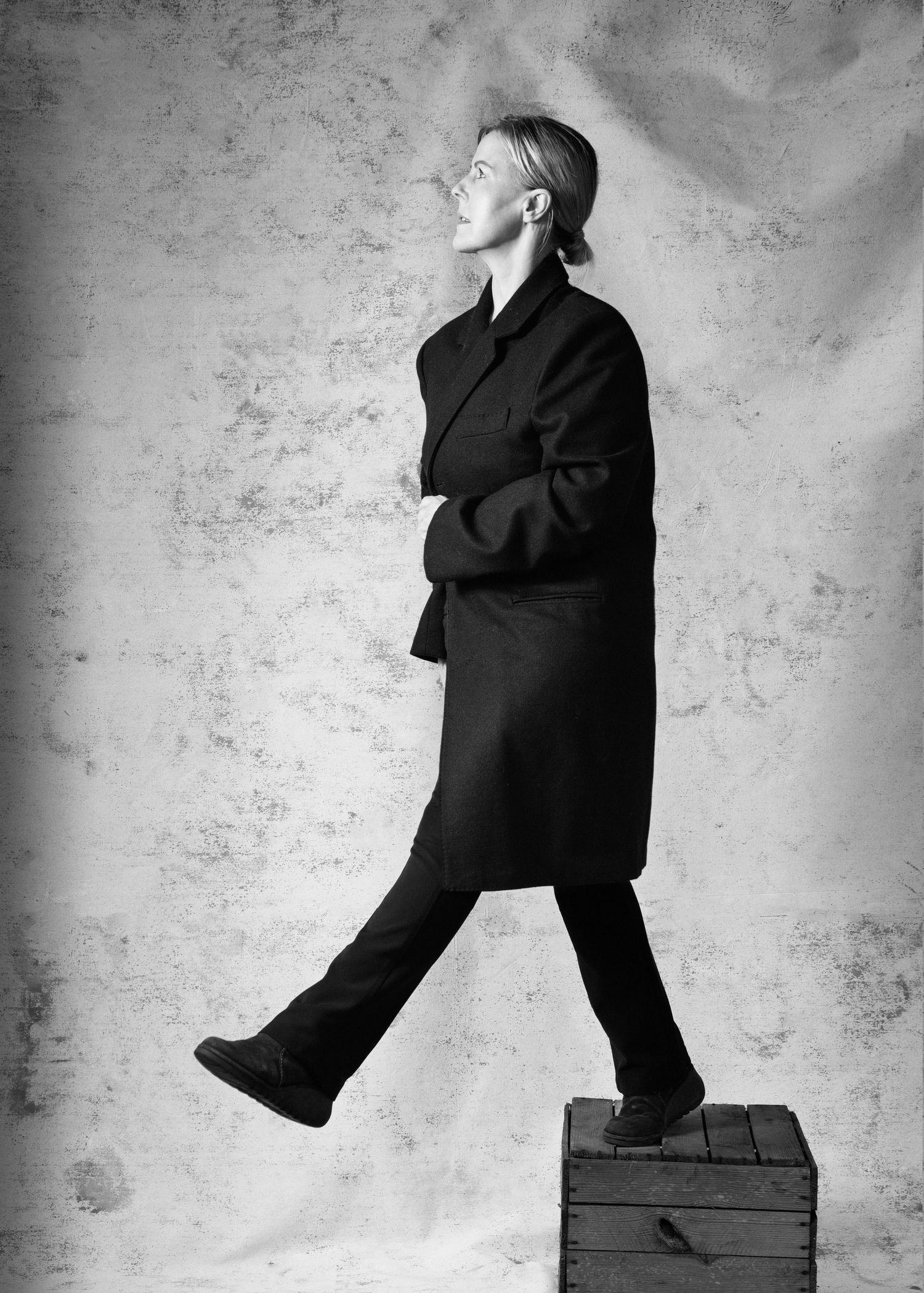 Black and white photo of a person in a dark oversized coat stepping off a wooden crate, looking upward.