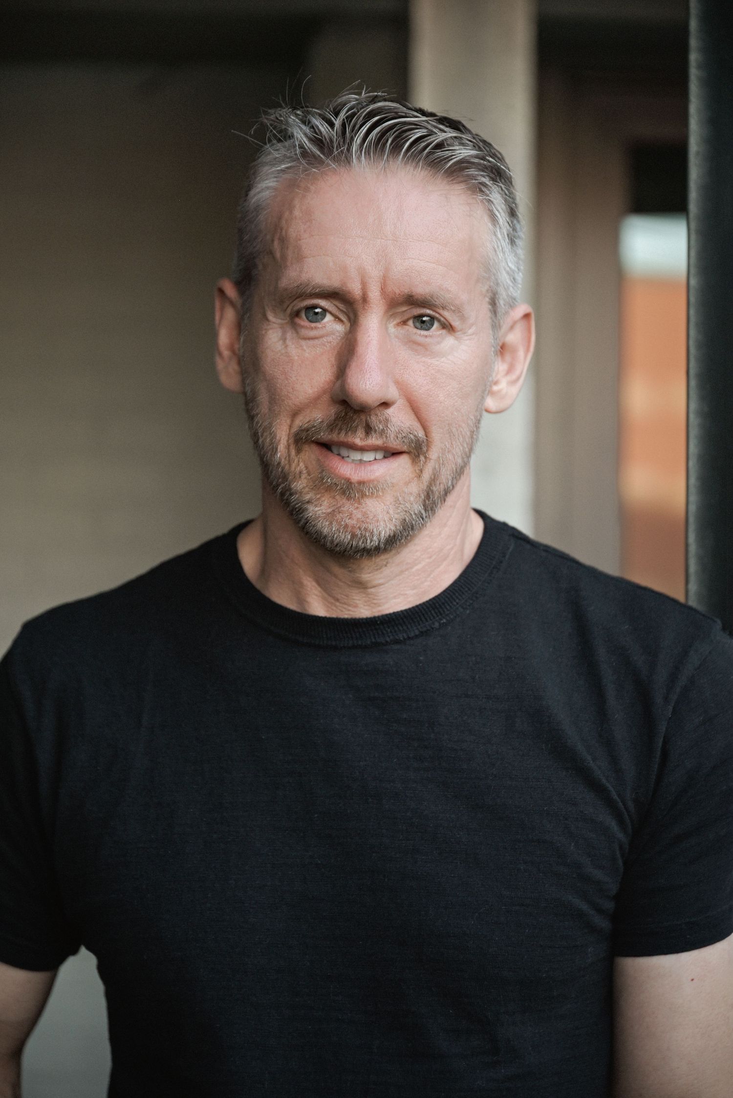 Middle-aged man with gray hair wearing a black t-shirt, smiling confidently in an outdoor urban setting.