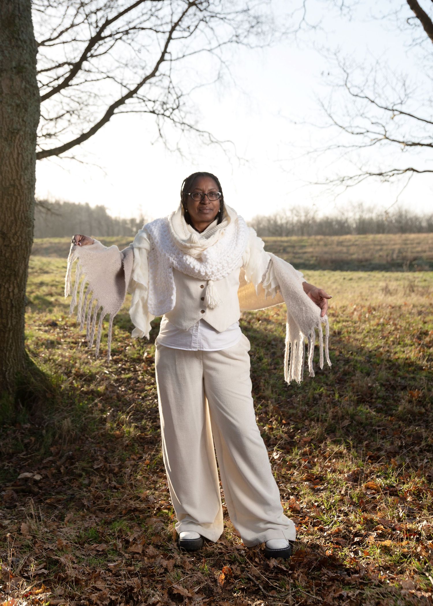 A person in white clothing stands in a wooded area with fallen autumn leaves on the ground during golden hour.