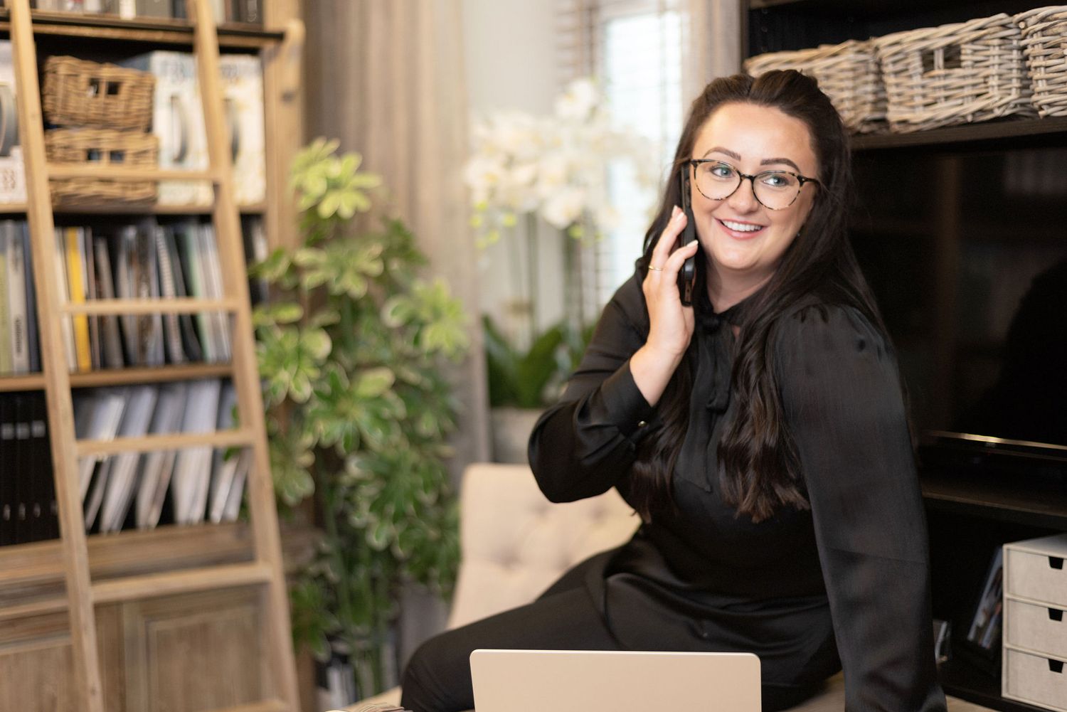 Professional taking phone call while seated in modern office with organized shelving and greenery.
