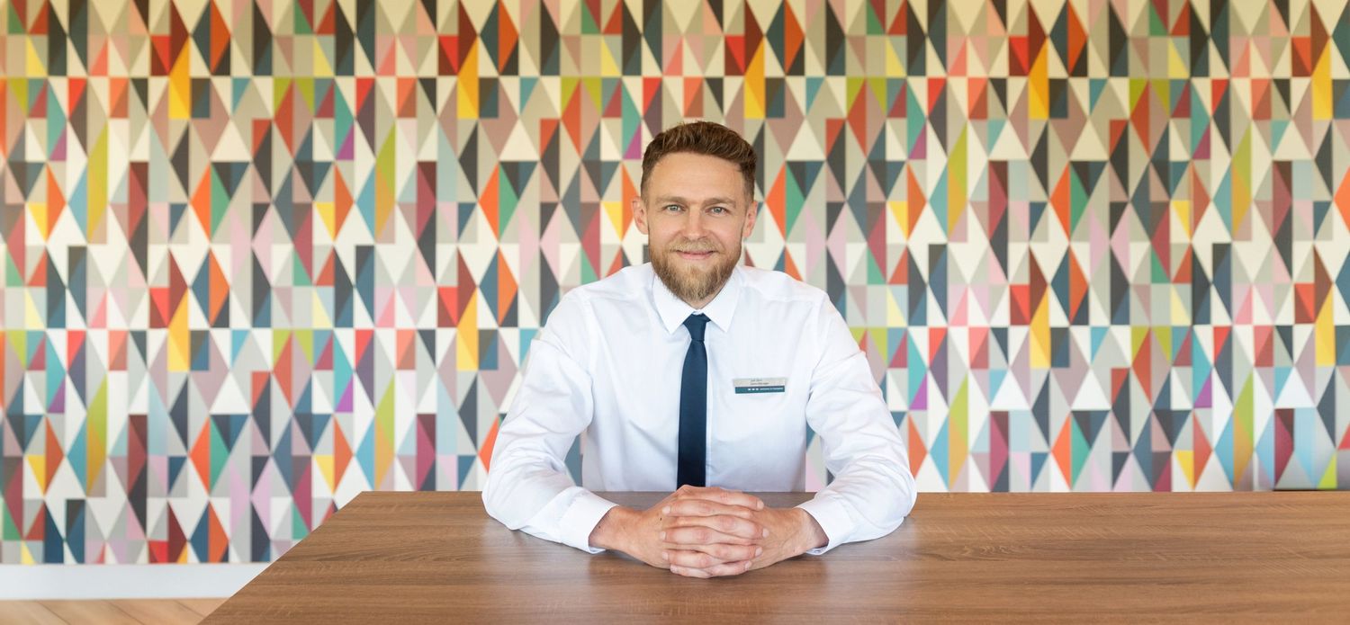 Professional in white dress shirt and tie sits at wooden desk against colourful geometric patterned wall.