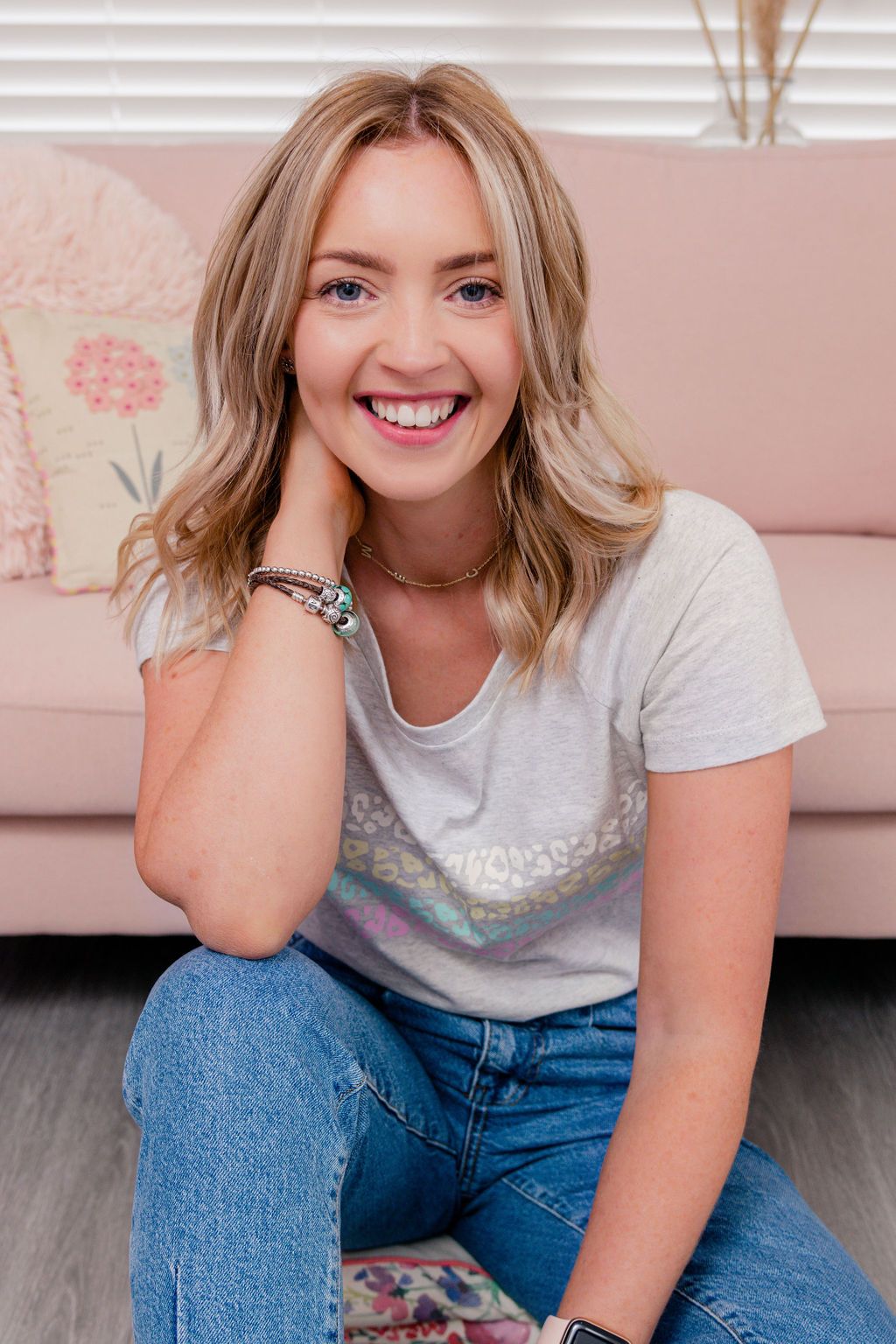 Smiling blonde woman in jeans and graphic tee sits on floor in front of pink sofa, wearing turquoise jewelry.