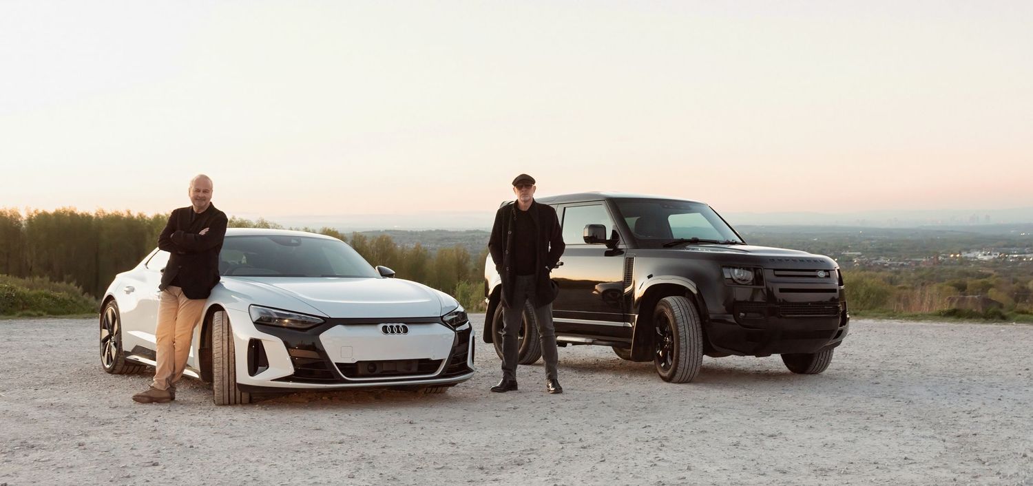 Two men stand beside a white Audi e-tron GT and black Land Rover Defender on a scenic overlook at dusk.