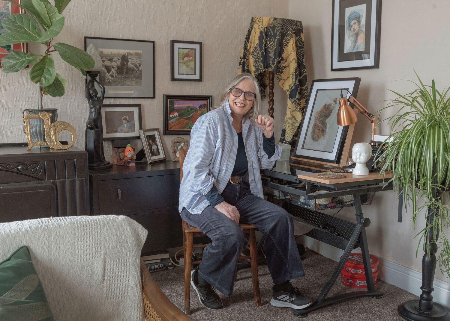 Woman with gray hair sits smiling at a desk surrounded by eclectic artwork and plants in a cozy, art-filled room.