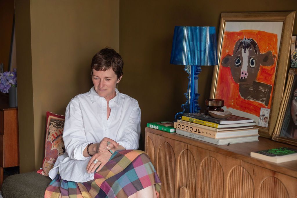 Woman in white shirt sits in armchair beside colorful artwork and blue lamp on wooden sideboard.