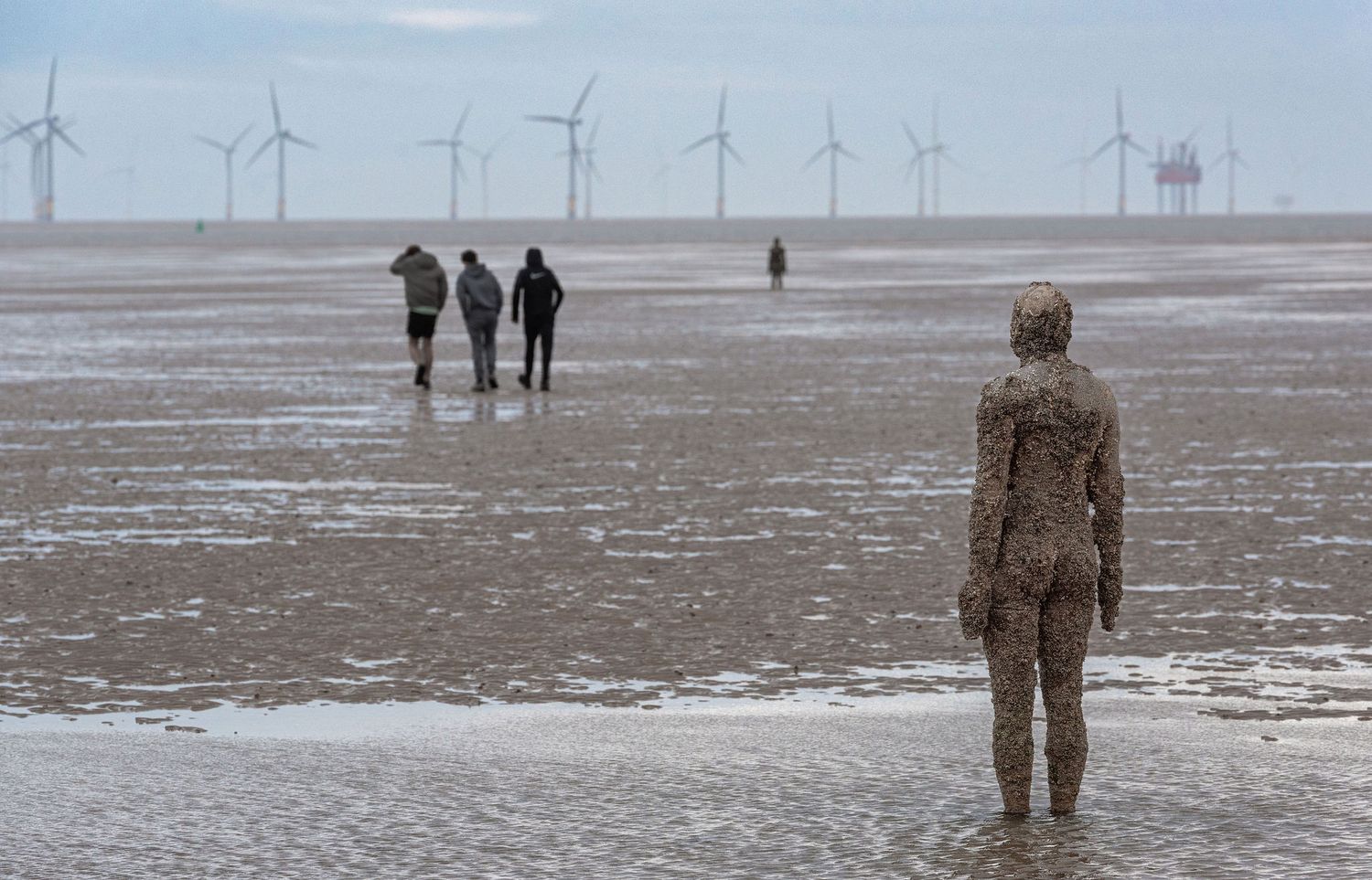 Iron Man statue on Crosby Beach with wind turbines and misty shoreline in background, Liverpool, England.