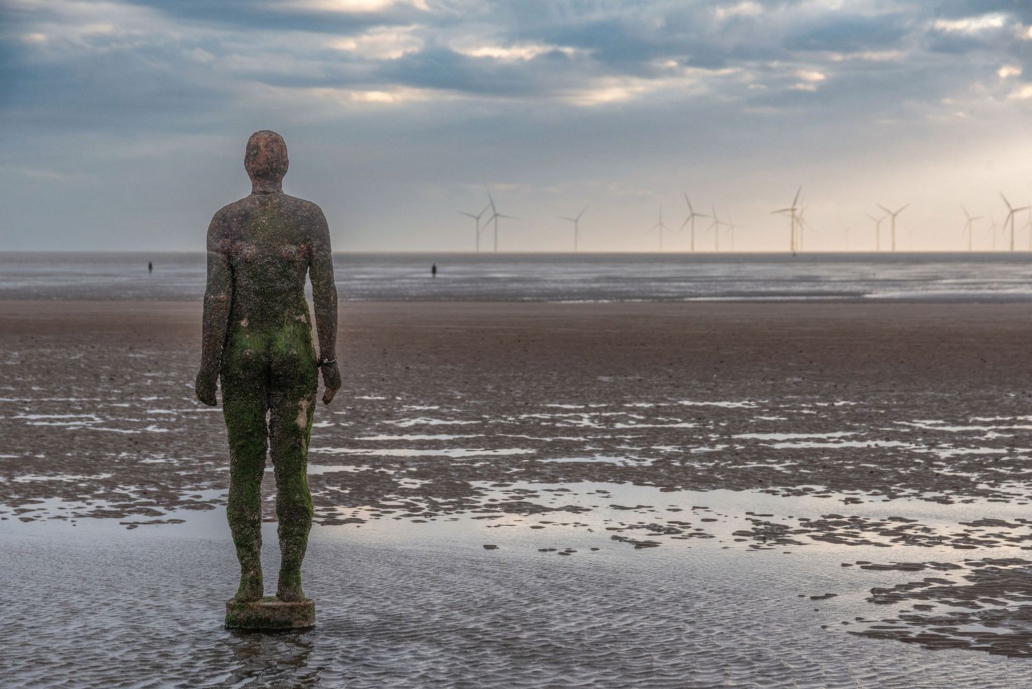Iron statue on a sandy beach at low tide with wind turbines and cloudy sky in the background.