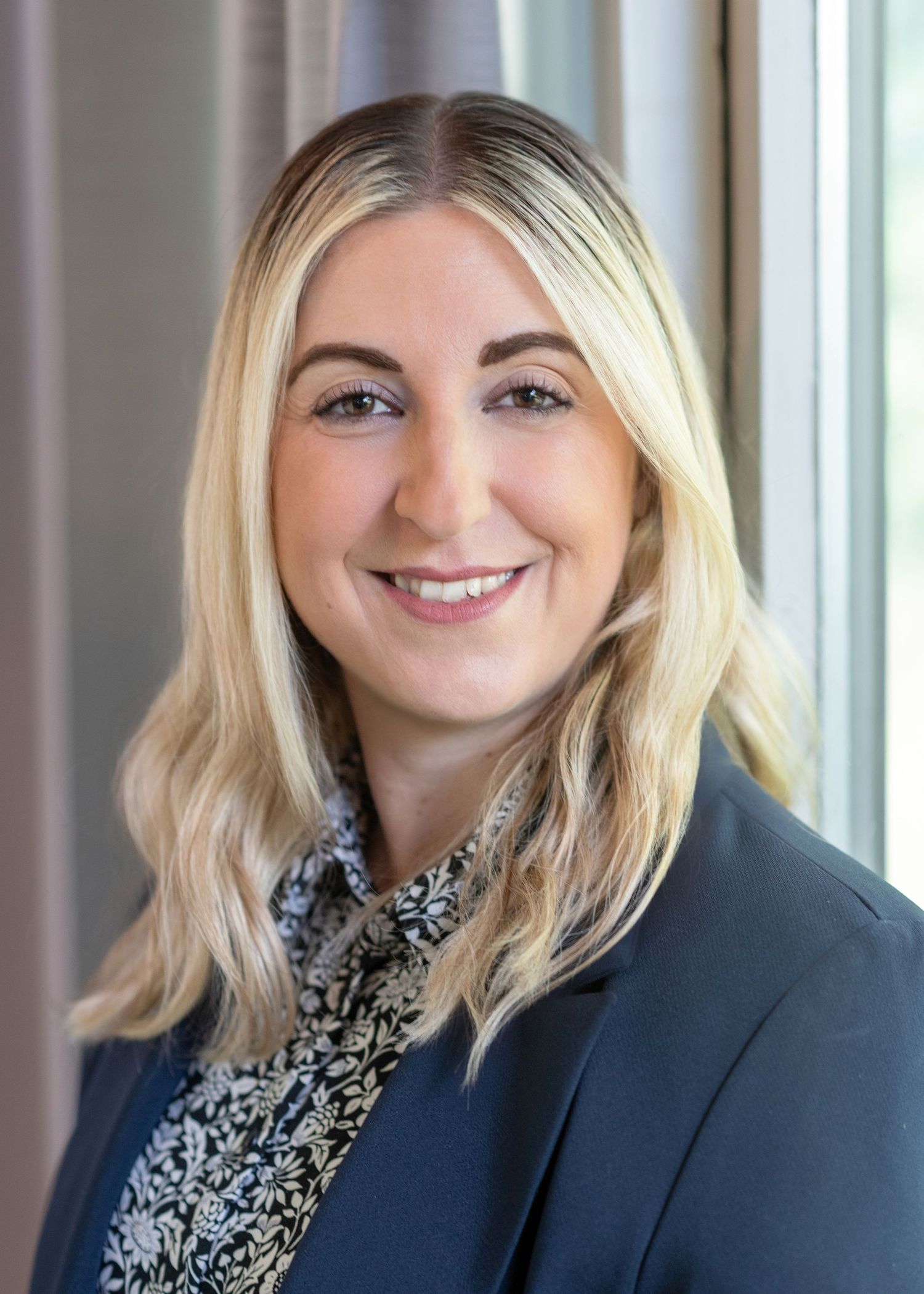 Professional headshot of a woman in a navy blazer and patterned blouse smiling by a window.