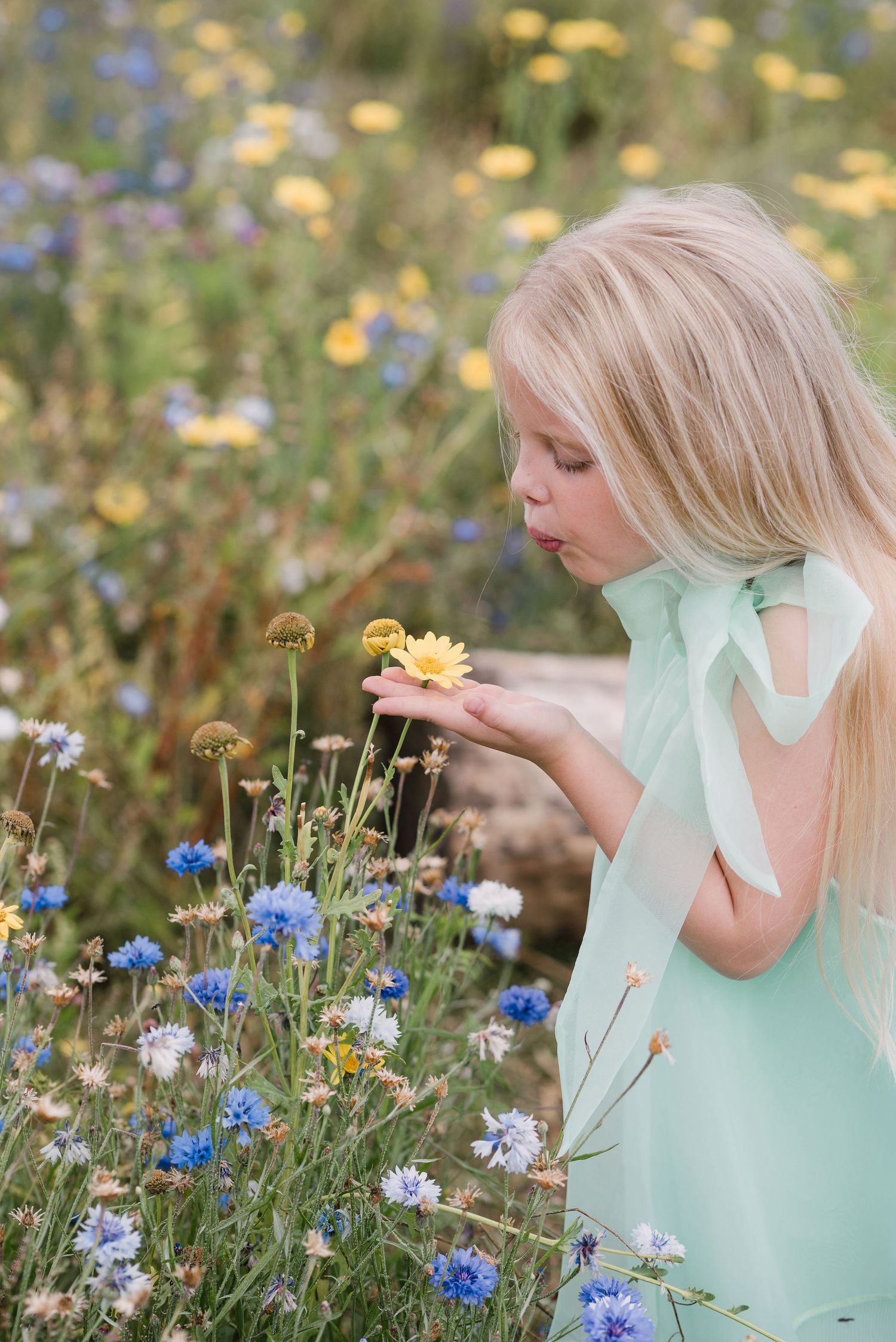 Someone with long blonde hair in a mint green dress smells wildflowers in a meadow of blue and yellow blooms.