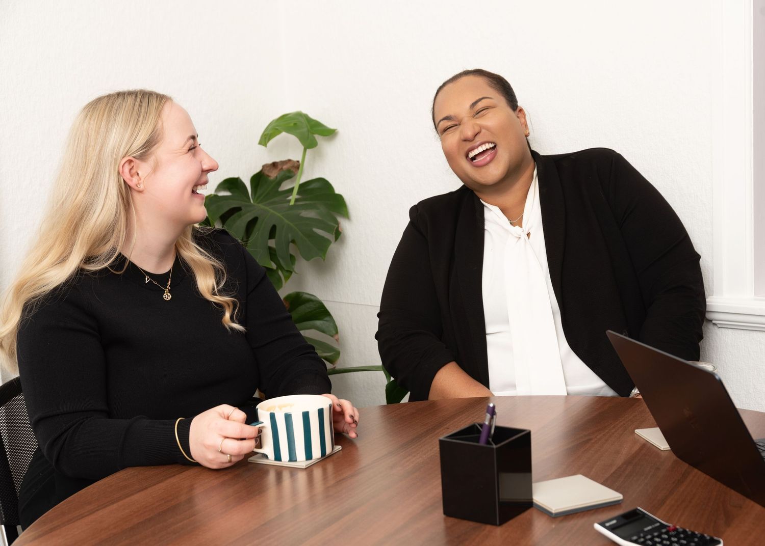 Two women laughing together at a office table, one holding a striped mug, with a laptop and plant in background.