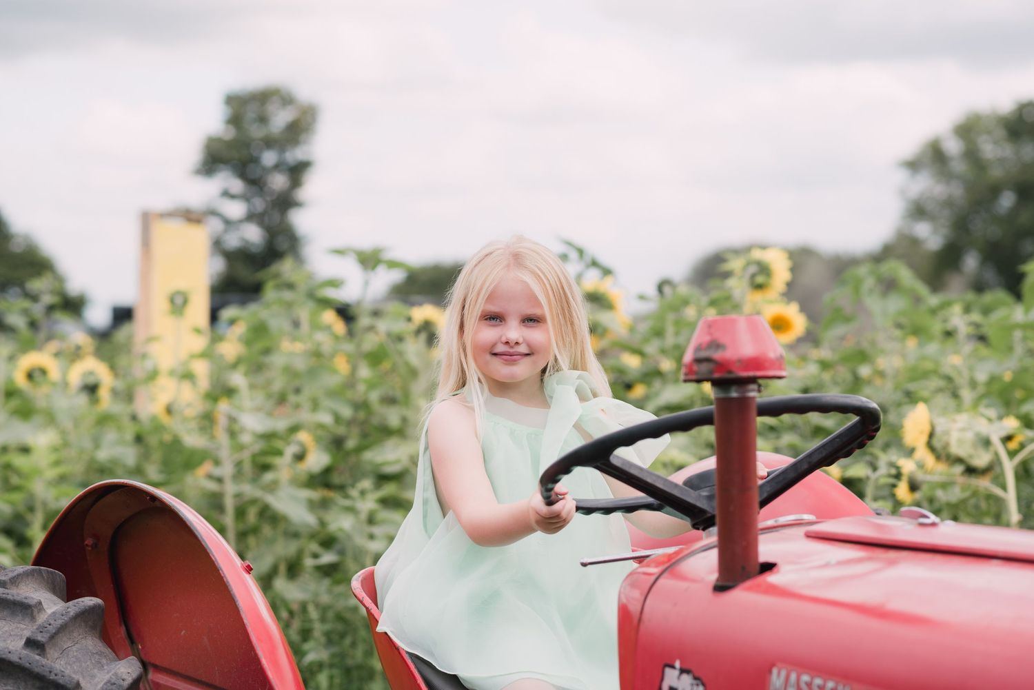 Young blonde girl in mint dress smiling on red vintage tractor surrounded by sunflowers in a farm field.