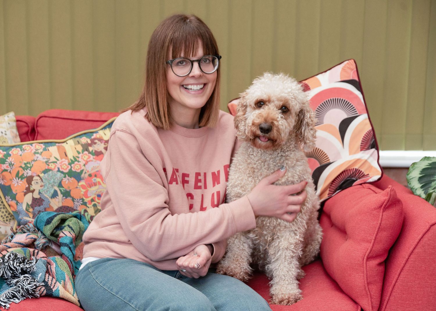 A small fluffy dog sits on a pink couch with someone wearing a pink sweater.