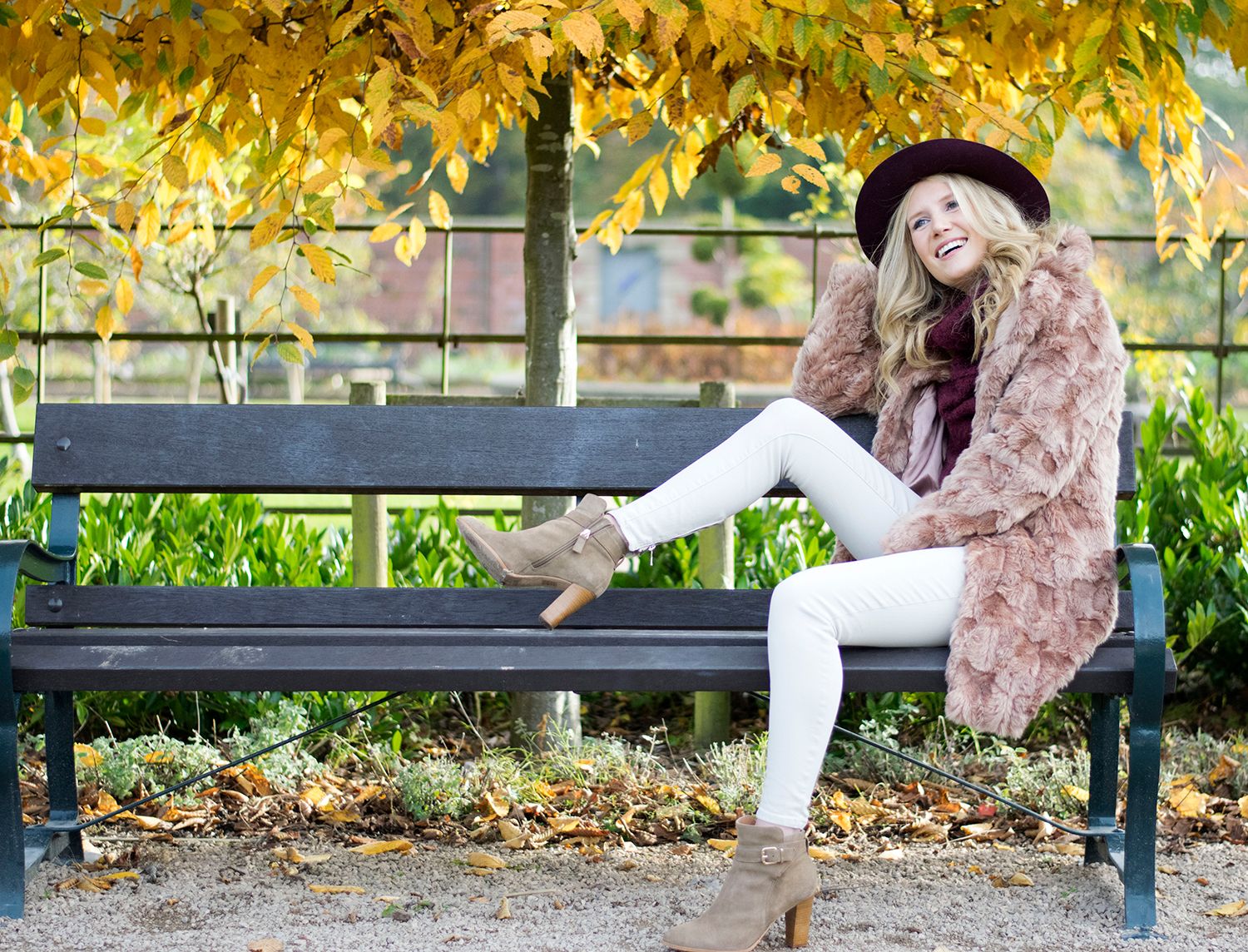Woman in pink fur coat and white jeans sitting on a park bench under golden autumn leaves.