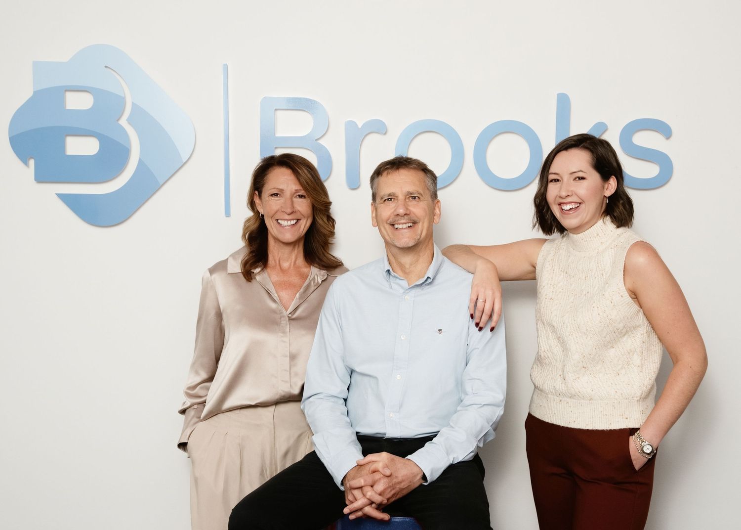 Three professionals smiling in front of a Brooks company logo sign in a modern office setting.