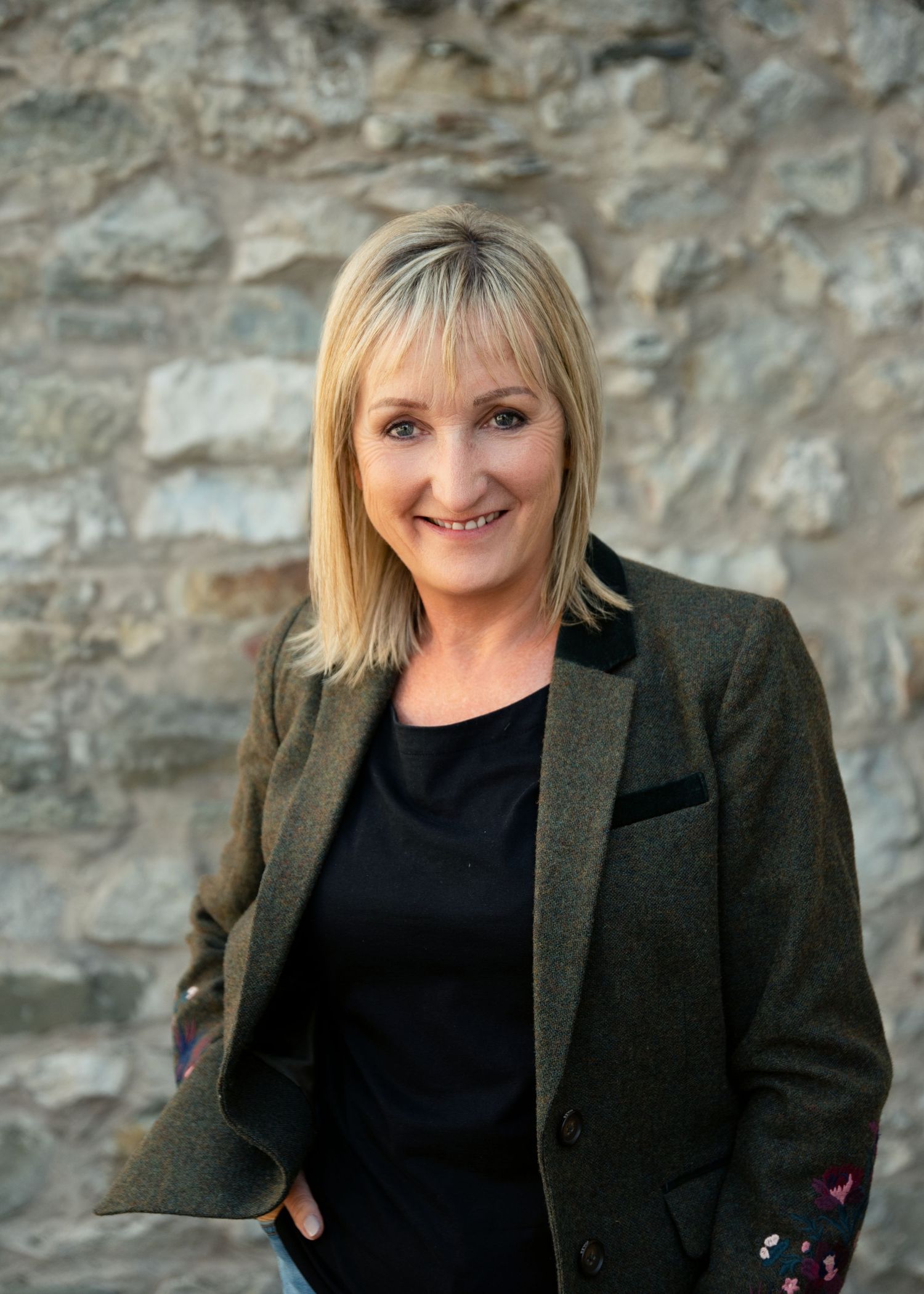 Smiling blonde woman in a dark blazer posing confidently against a rustic stone wall background.