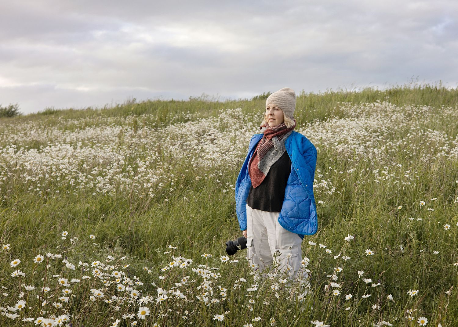 Woman in blue jacket and hat stands among white wildflowers in a lush green meadow under cloudy skies.