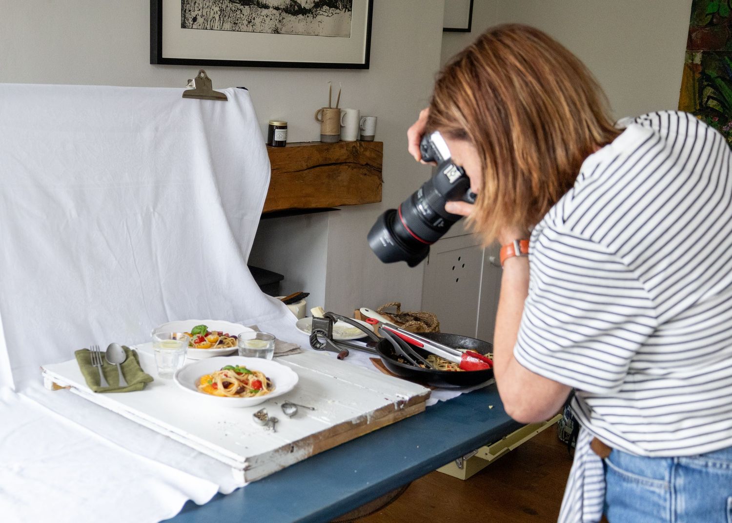Woman photographer taking close-up shots of plated food dishes on a white backdrop table in a home studio setting.