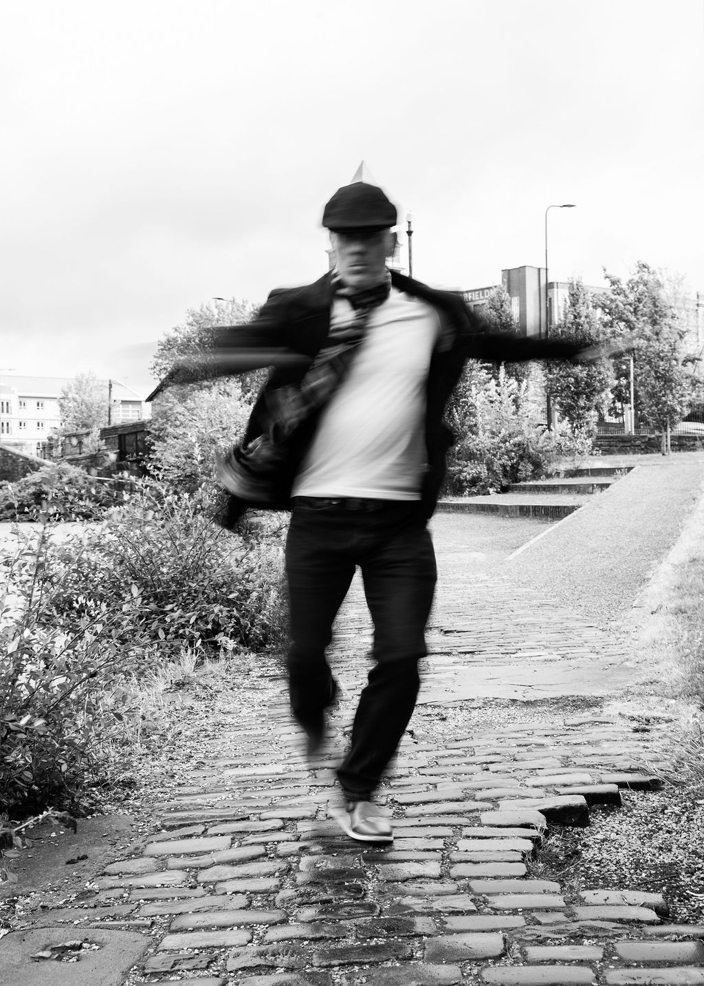 Black and white photo of a man in motion blur, wearing a cap and jacket, walking on a cobblestone path.